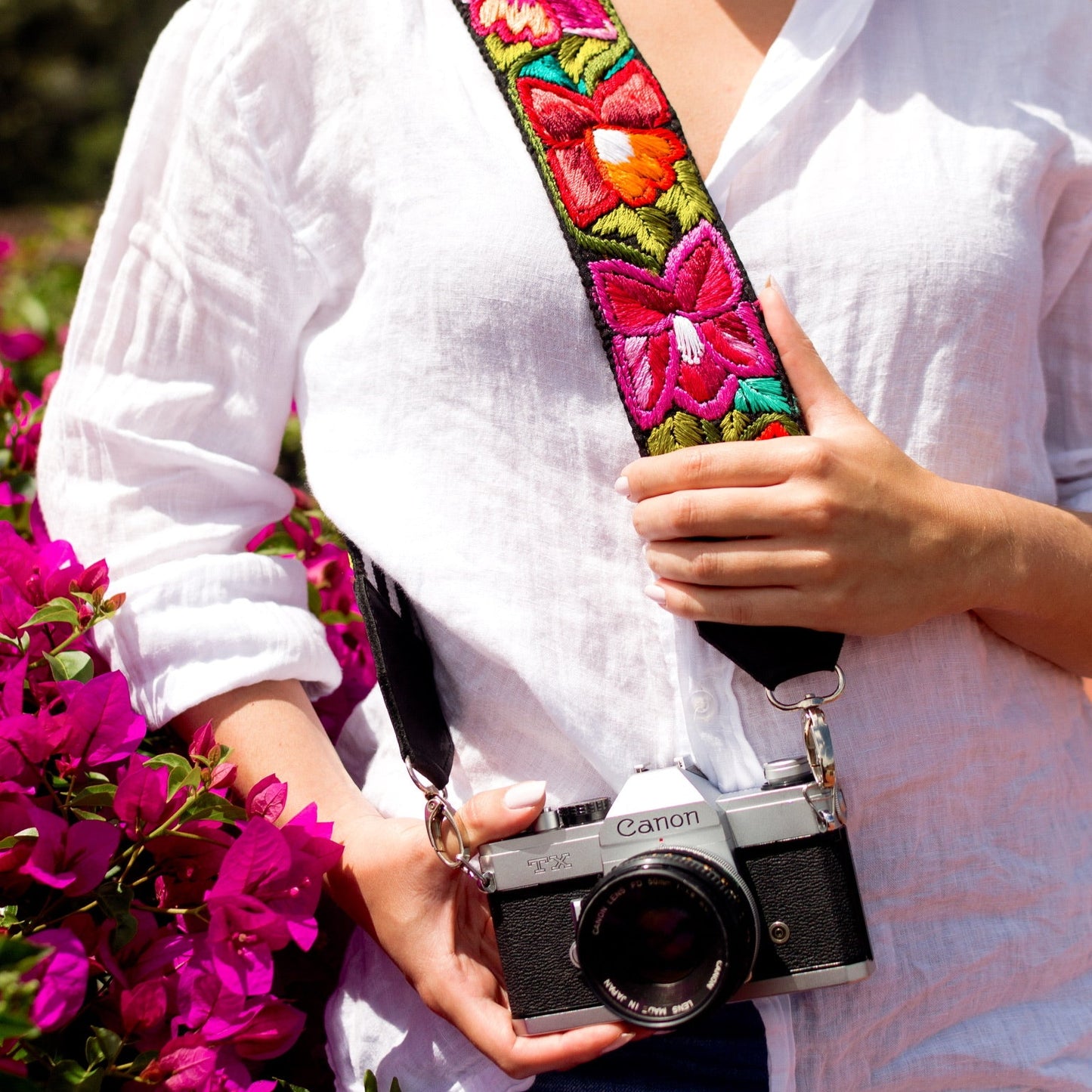Person holding a camera with a colorful floral strap, surrounded by flowers outdoors.