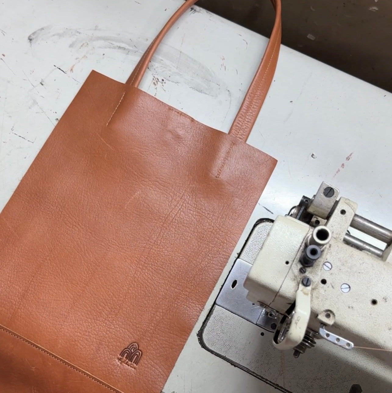 Brown leather tote bag on a sewing machine with a visible brand logo.
