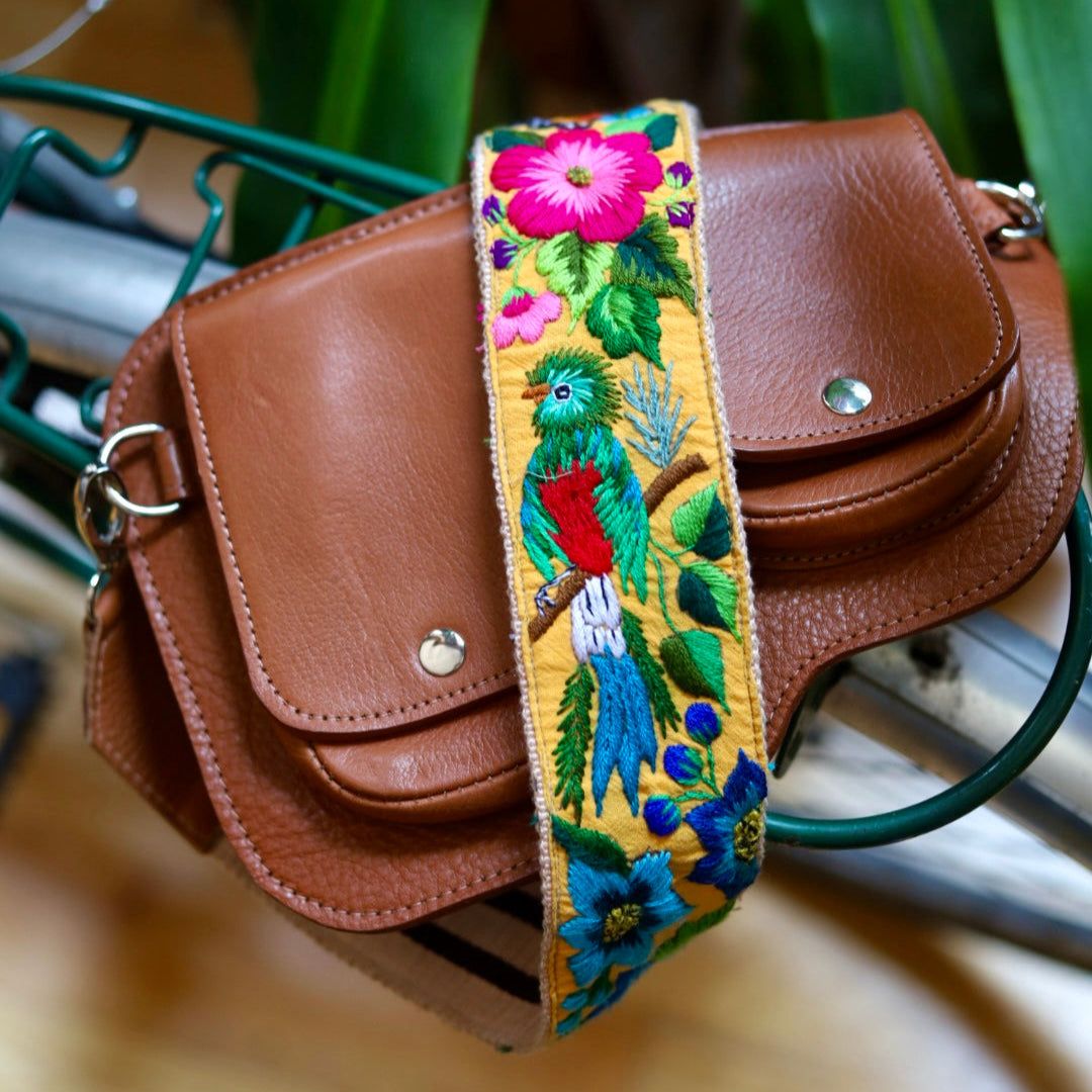 Brown leather bag with a colorful embroidered strap featuring birds and flowers.