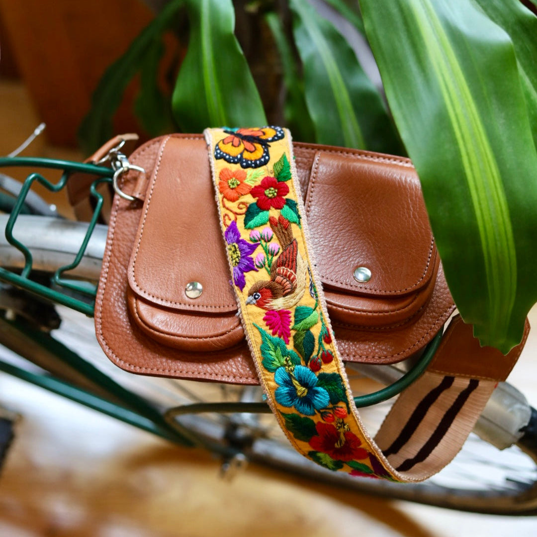 Brown leather bag with a colorful floral strap on a bicycle, surrounded by green plants.