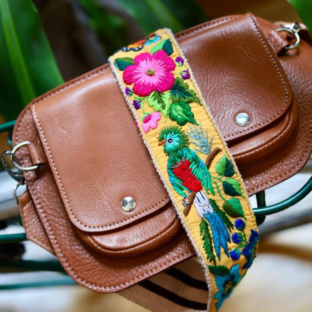 Brown leather bag with a colorful embroidered strap featuring birds and flowers.
