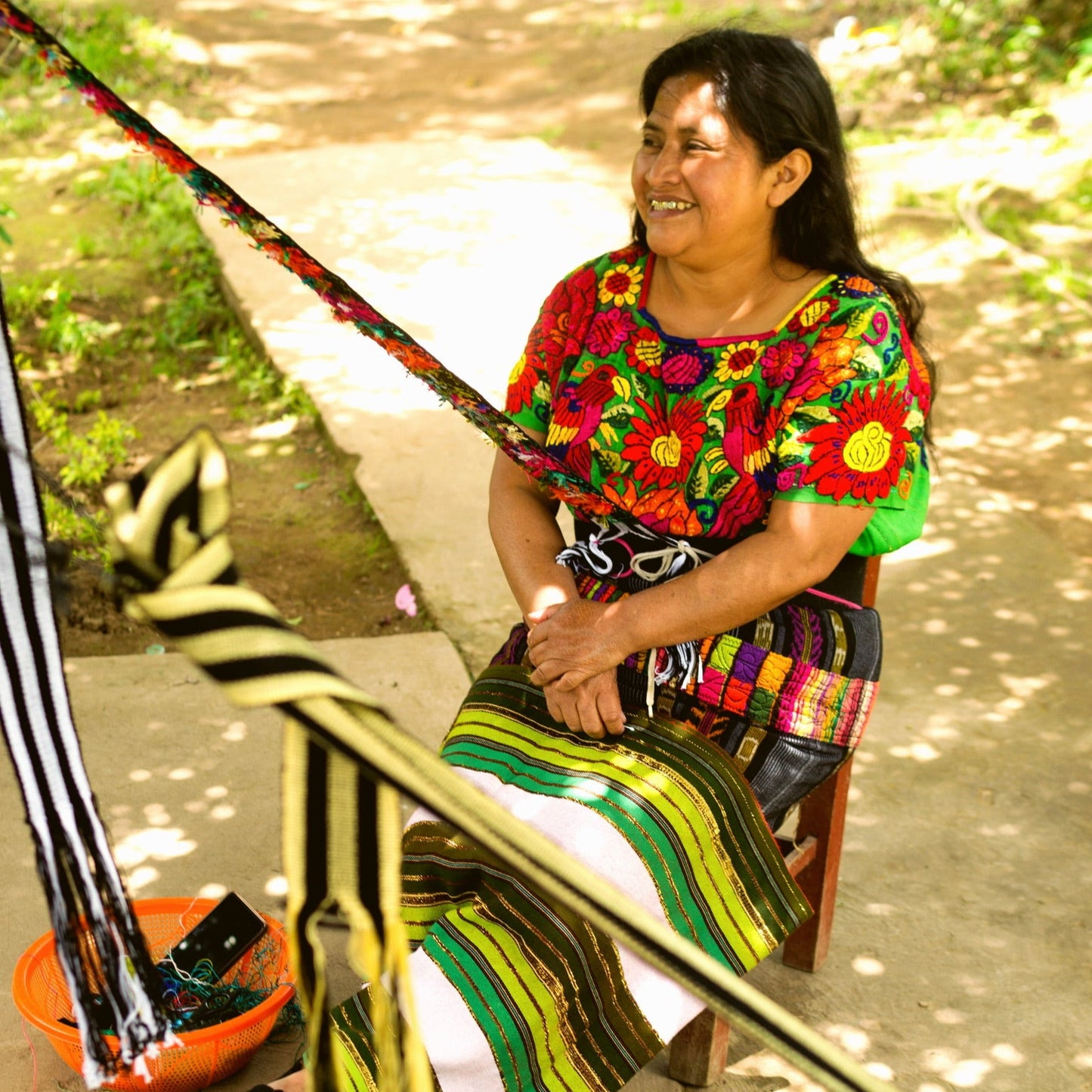 Woman in colorful traditional clothing sitting and working with a loom outdoors.
