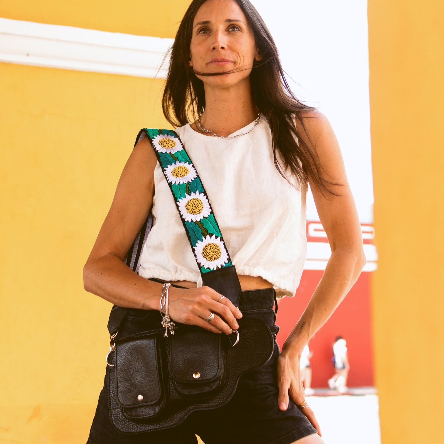 Woman holding a black bag with a colorful strap against a yellow wall.