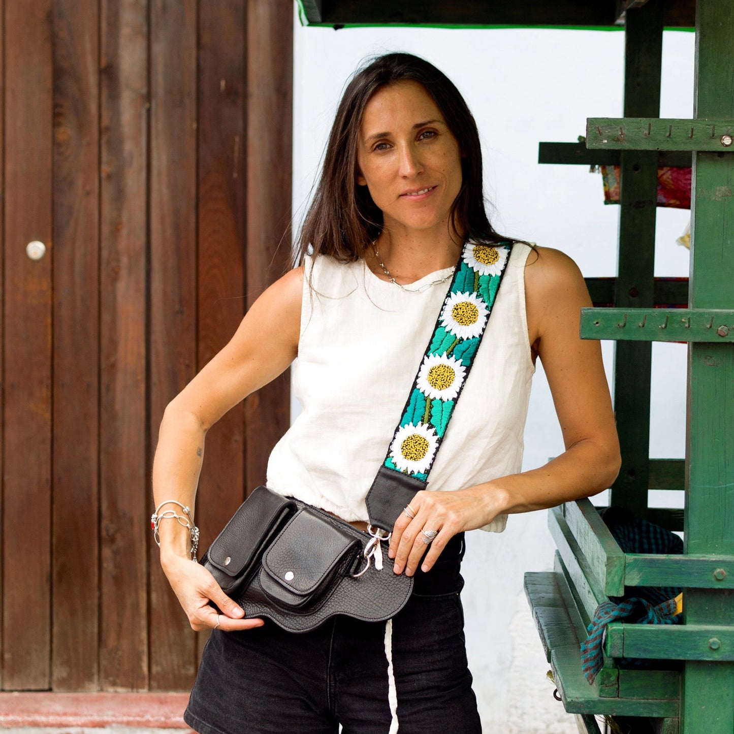 Woman holding a black bag with a colorful strap in front of a green structure.