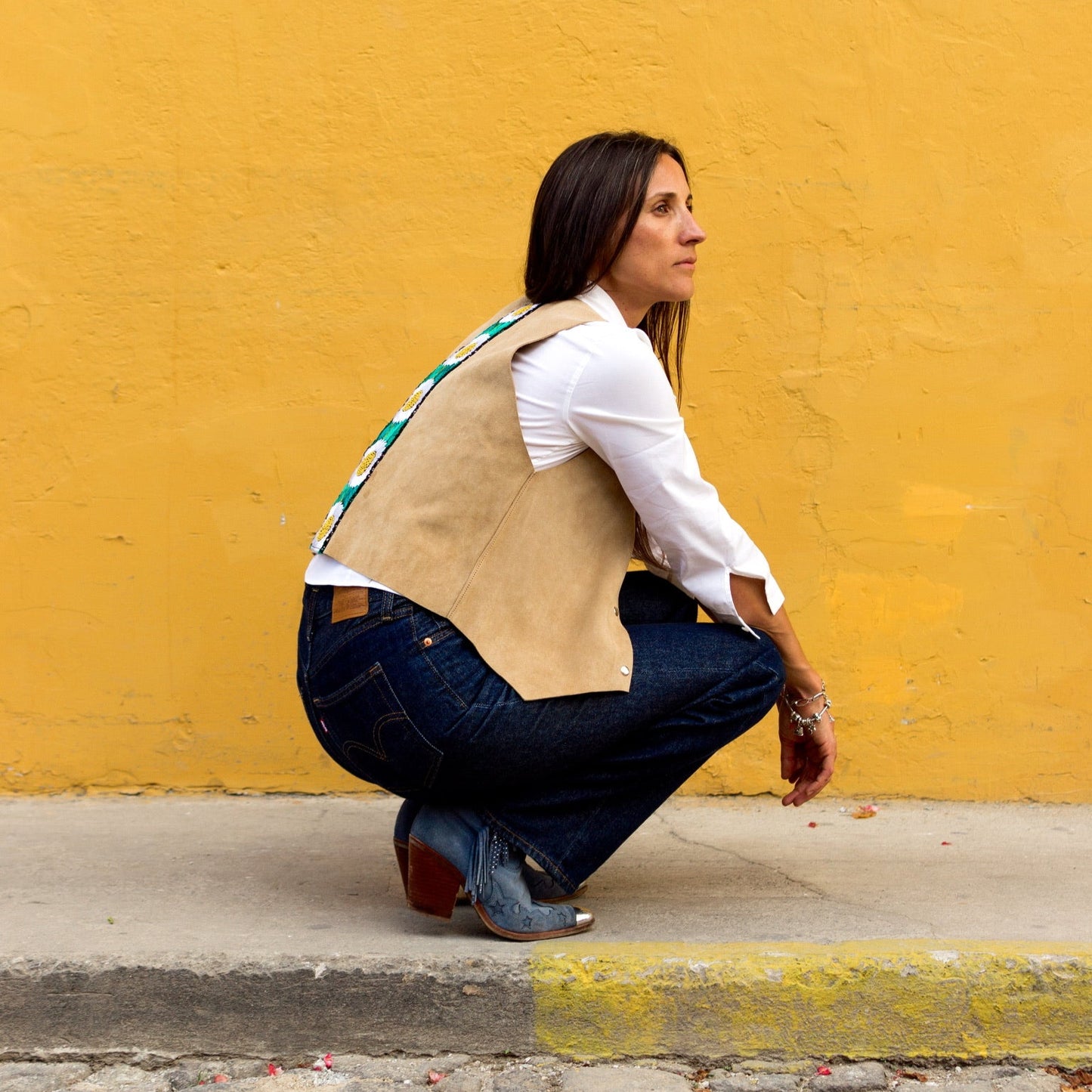 Woman in a beige vest and white shirt crouching against a yellow wall.