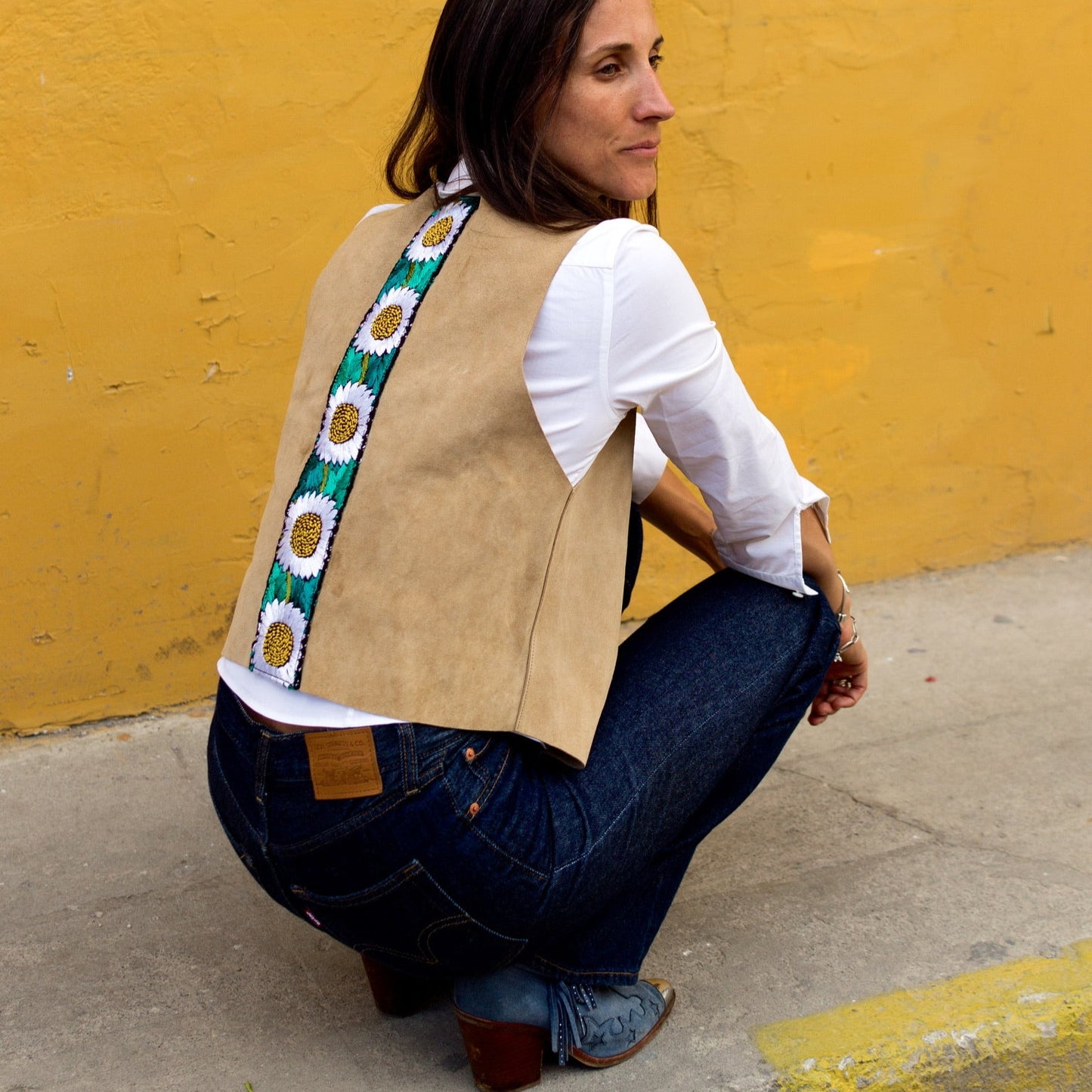 Woman wearing a tan vest with a colorful belt against a yellow wall.