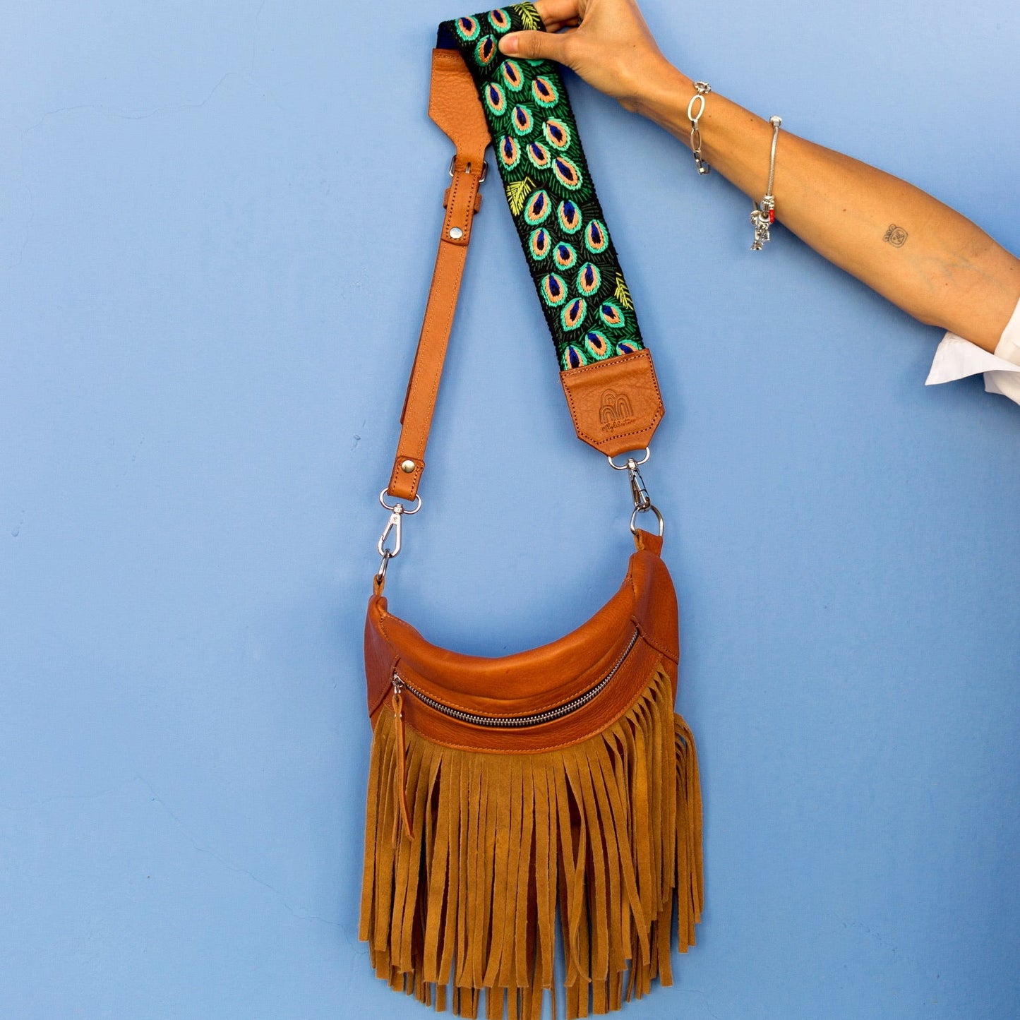 Brown leather handbag with peacock feather strap against a blue background