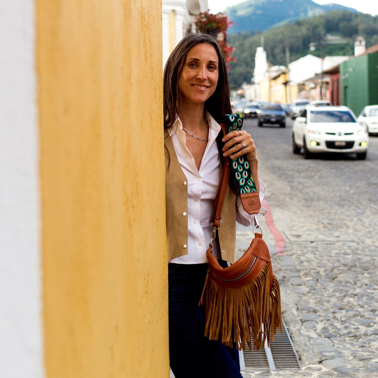 Woman holding a coffee cup and a handbag, standing in a street with mountains in the background.