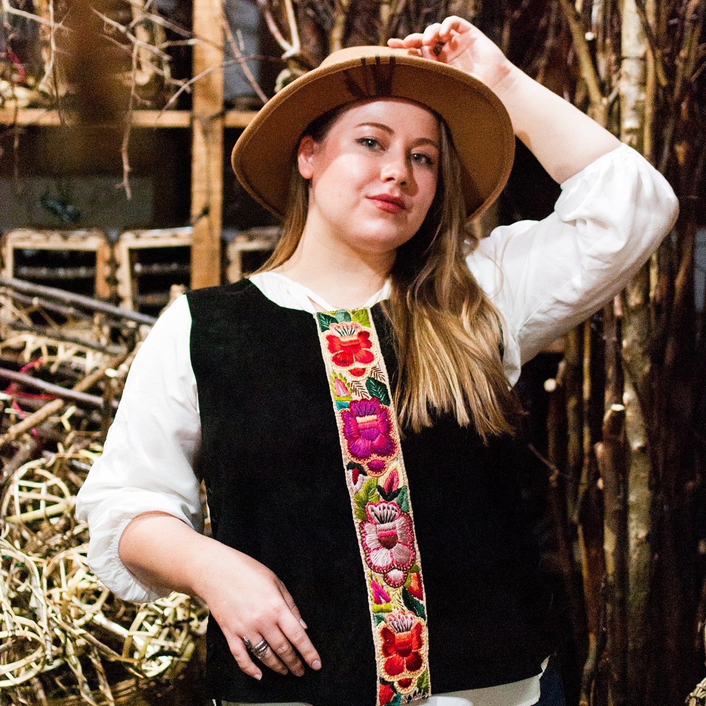 Woman wearing a black vest with floral patterns and a wide-brimmed hat in a rustic setting.