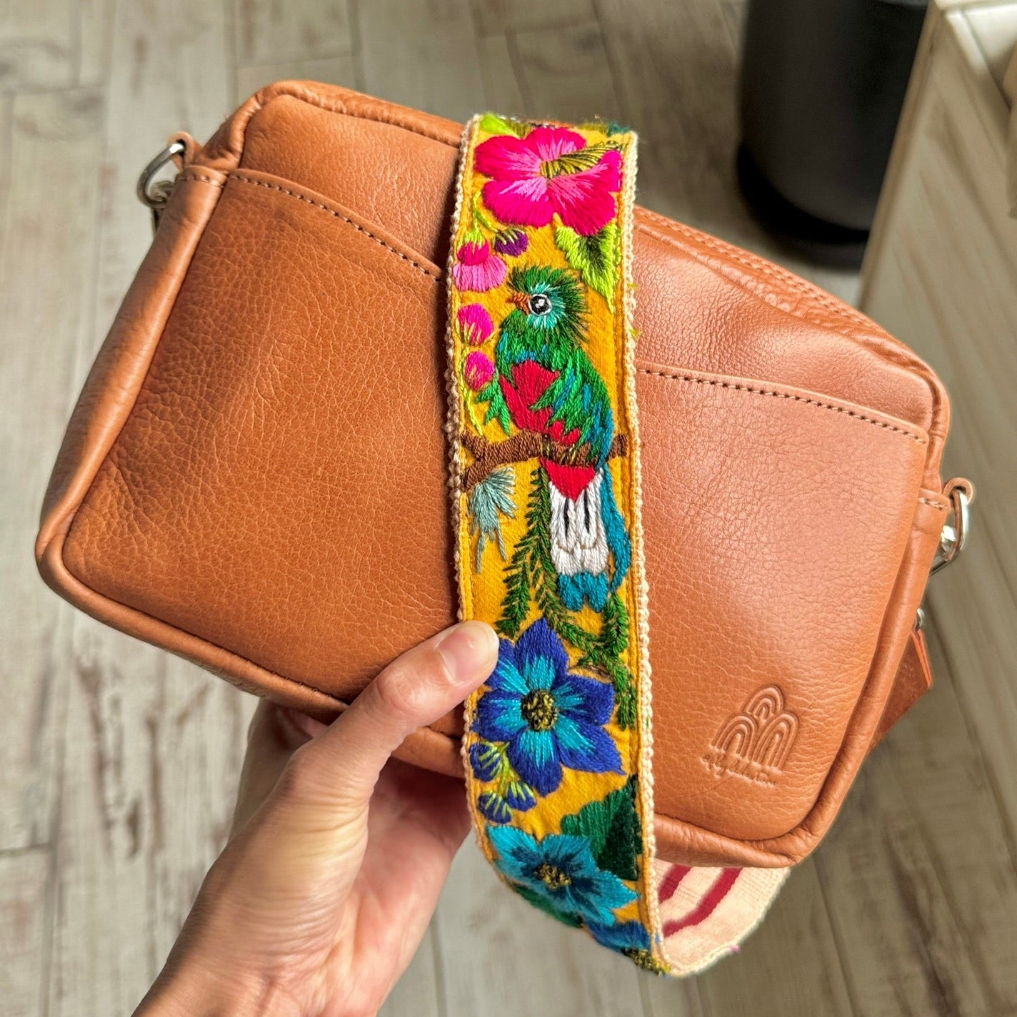 A tan leather crossbody bag with a colorful floral and parrot-patterned fabric strap, held by a hand against a light wood floor background.