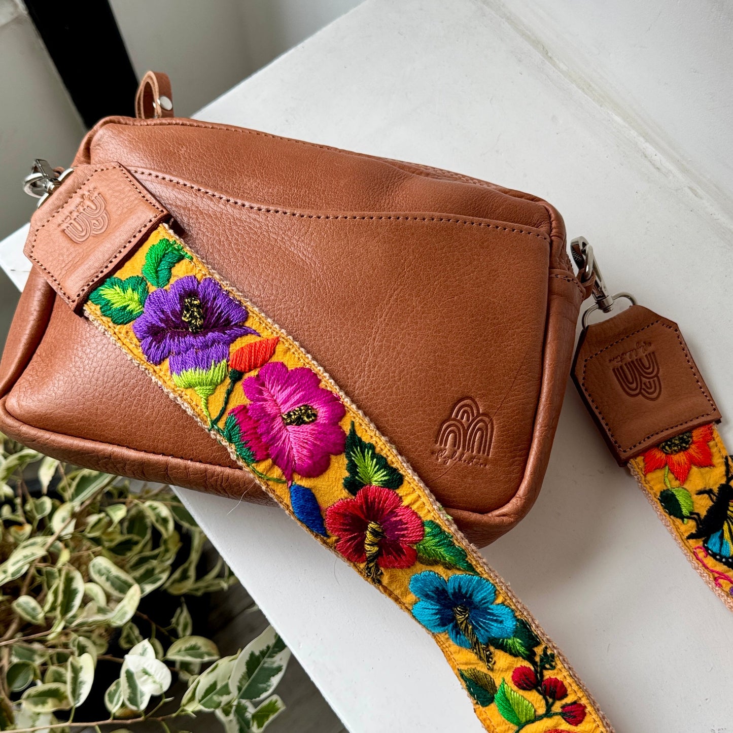 A brown leather crossbody bag with a wide, vibrant, hand-embroidered floral strap, resting on a white table next to a potted plant.