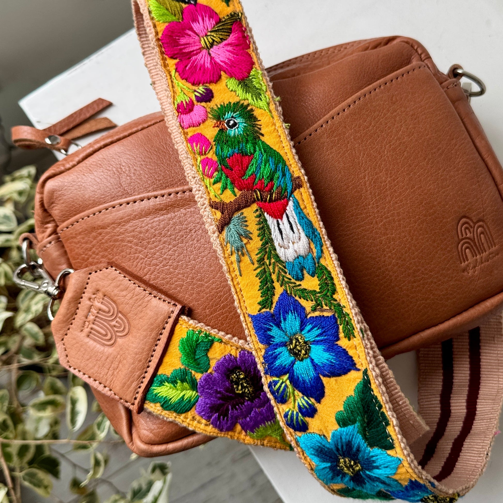 A brown leather crossbody bag with a wide, vibrant embroidered floral and bird shoulder strap, displayed on a white surface.