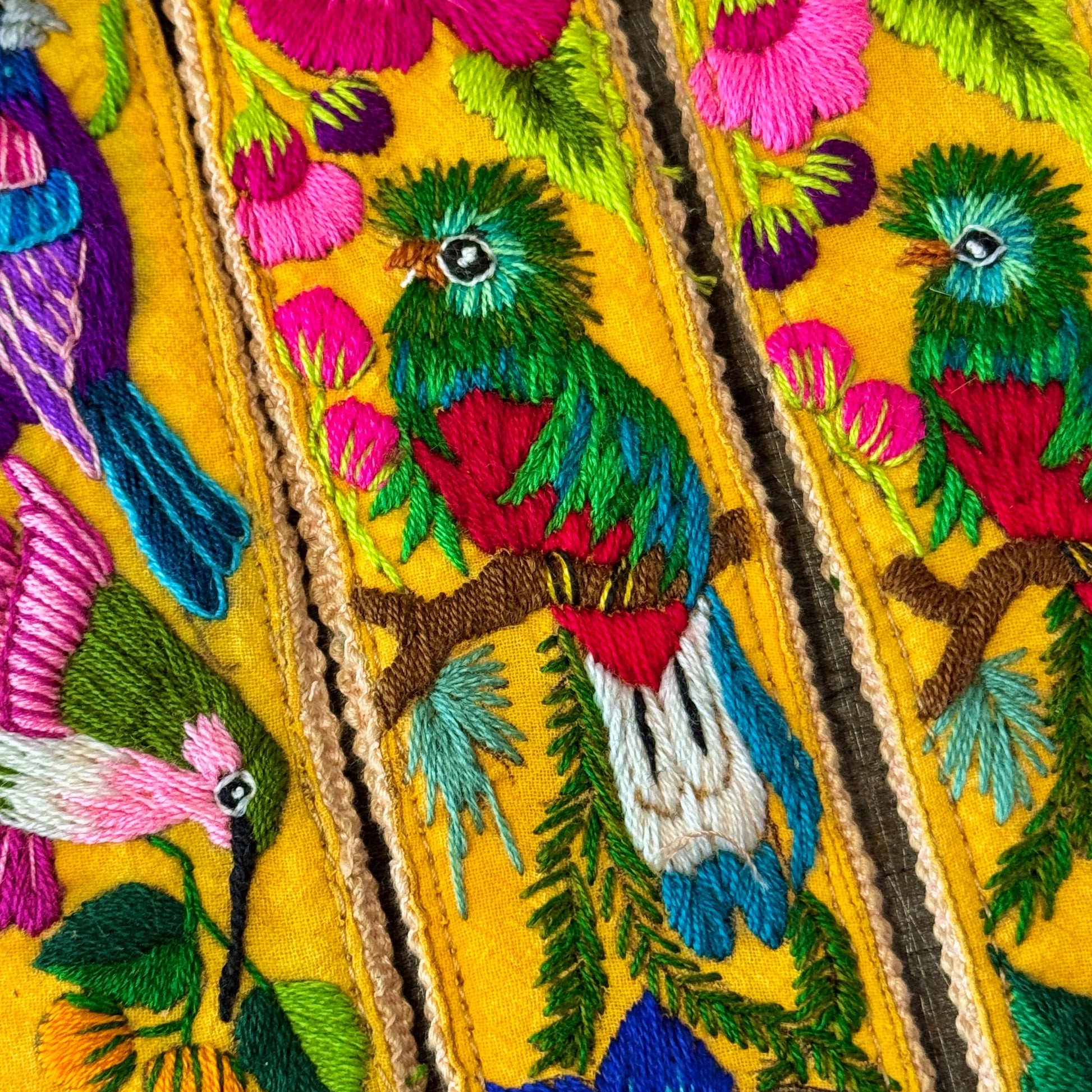 Close-up of a vibrant yellow embroidered textile featuring colorful birds and flowers, including pink hibiscus, blue flowers, and green birds with red accents.