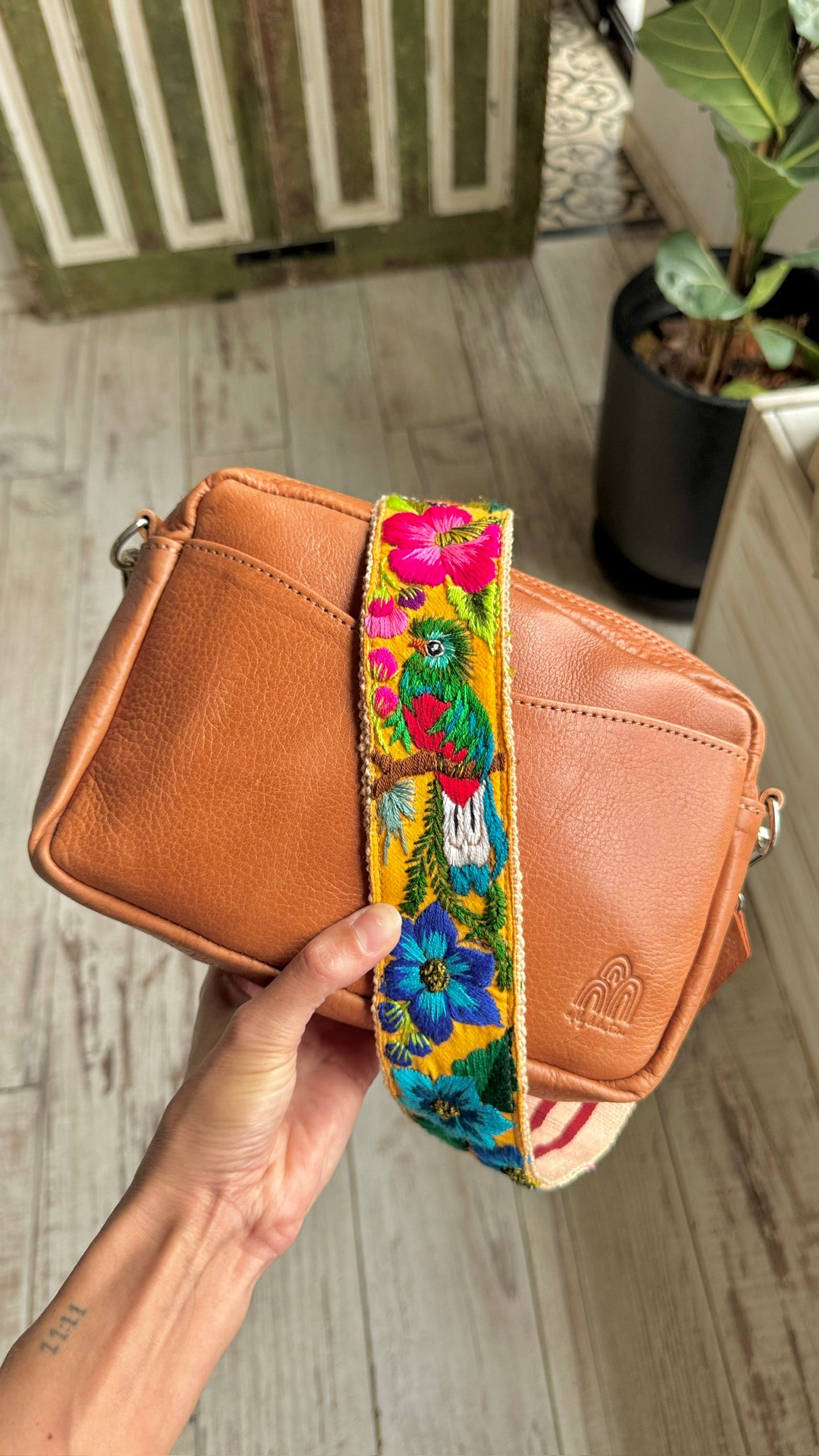 A tan leather crossbody bag with a colorful floral and parrot-patterned fabric strap, held by a hand against a light wood floor background.