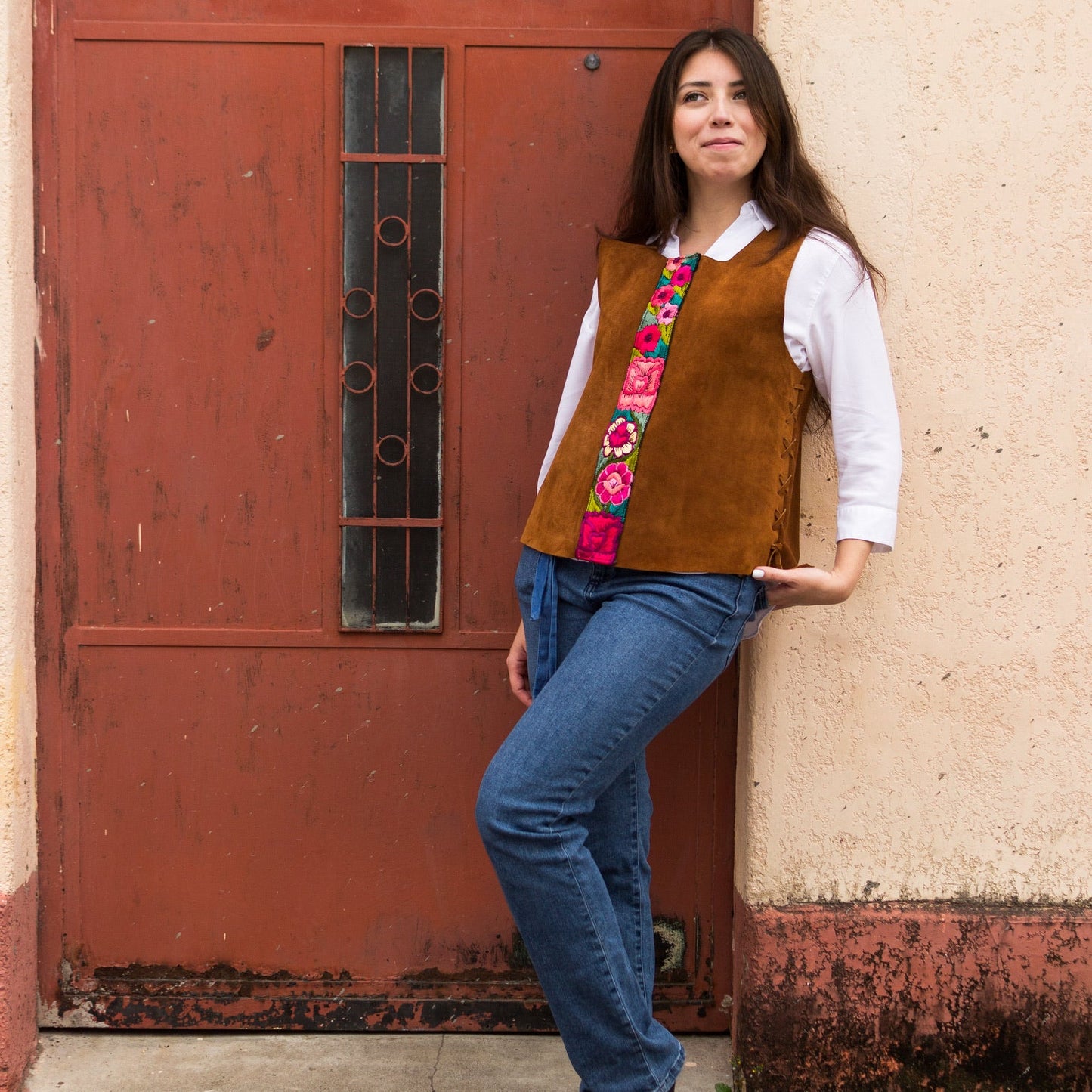 Woman wearing a brown vest with floral patterns leaning against a red door.