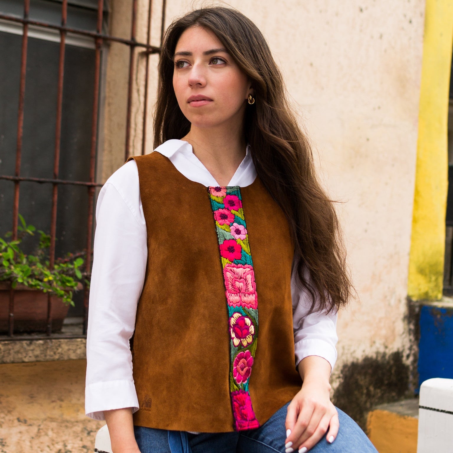 Woman wearing a brown vest with colorful floral patterns sitting against a wall.
