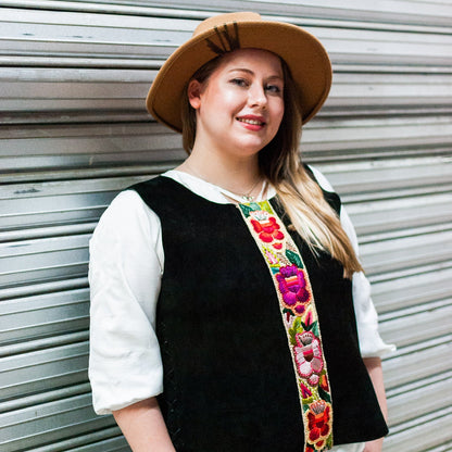 Woman wearing a black vest with colorful floral embroidery and a beige hat against a metallic background