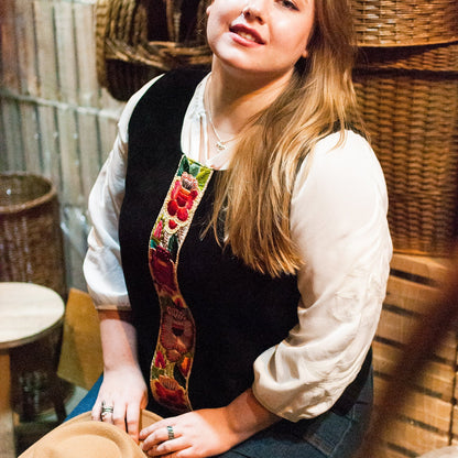 Woman sitting among wicker baskets wearing a colorful belt and white shirt.