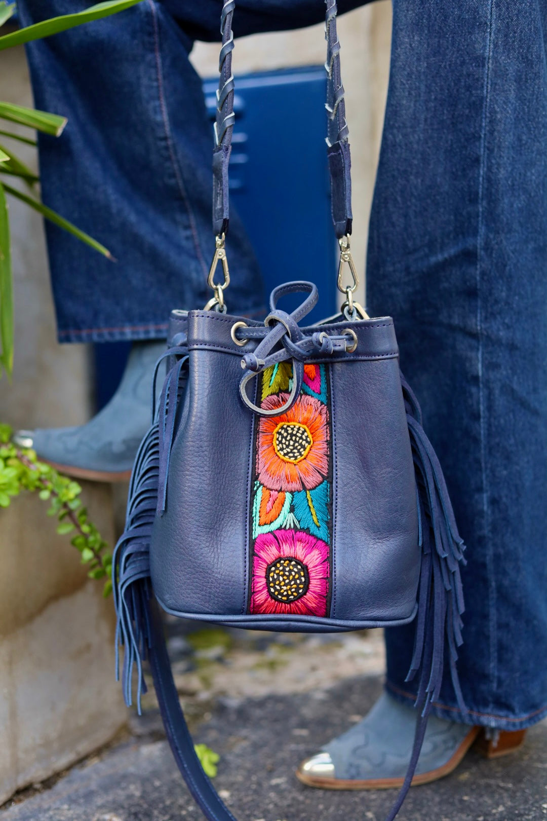 Blue handbag with floral designs and fringes held by a person wearing blue jeans and shoes.