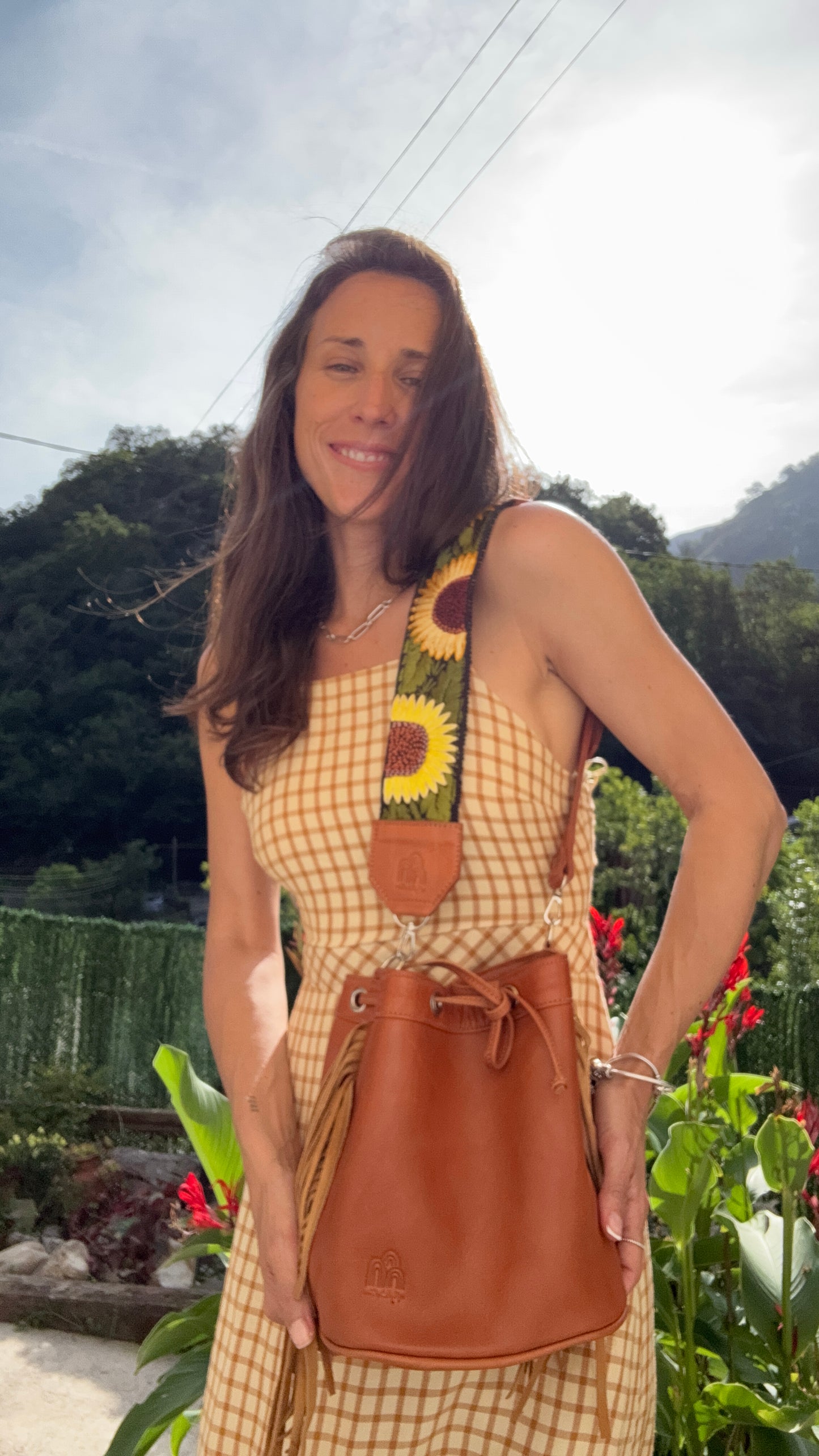 A woman outdoors wearing a yellow and white checkered dress and carrying a brown leather bucket bag with a sunflower-patterned strap.
