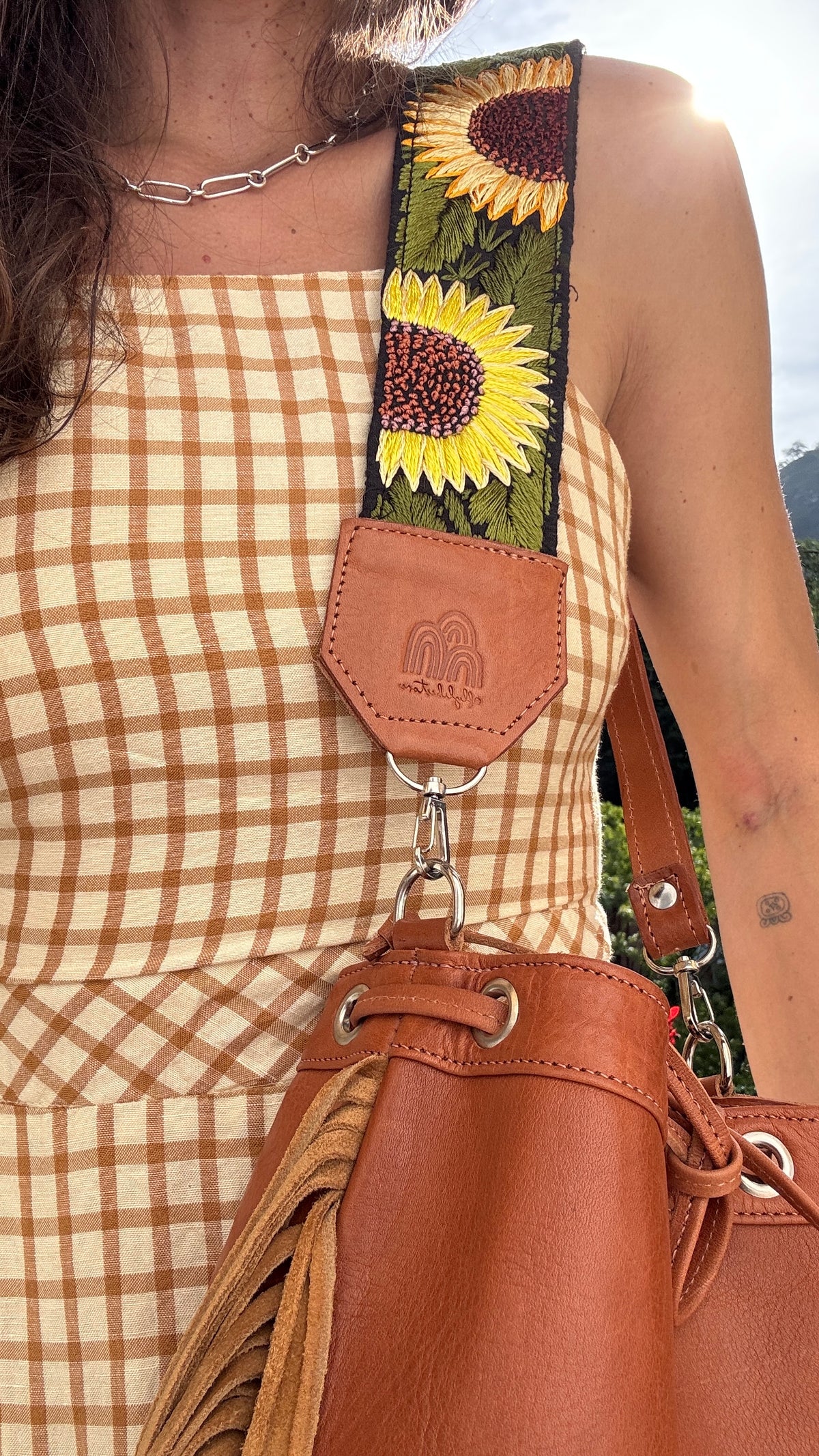 A close-up of a person wearing a brown and beige gingham dress and carrying a brown leather shoulder bag with a wide decorative strap featuring embroidered sunflowers.