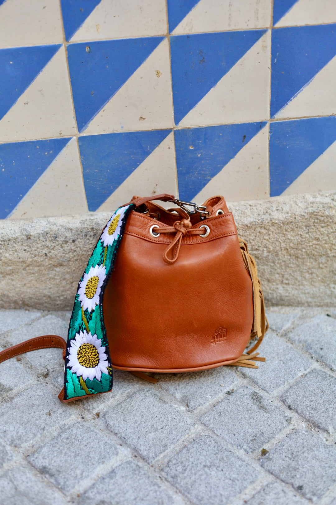 A brown leather bucket bag with a drawstring closure and a wide, colorful floral shoulder strap, resting on a stone pavement in front of a blue and beige tiled wall.