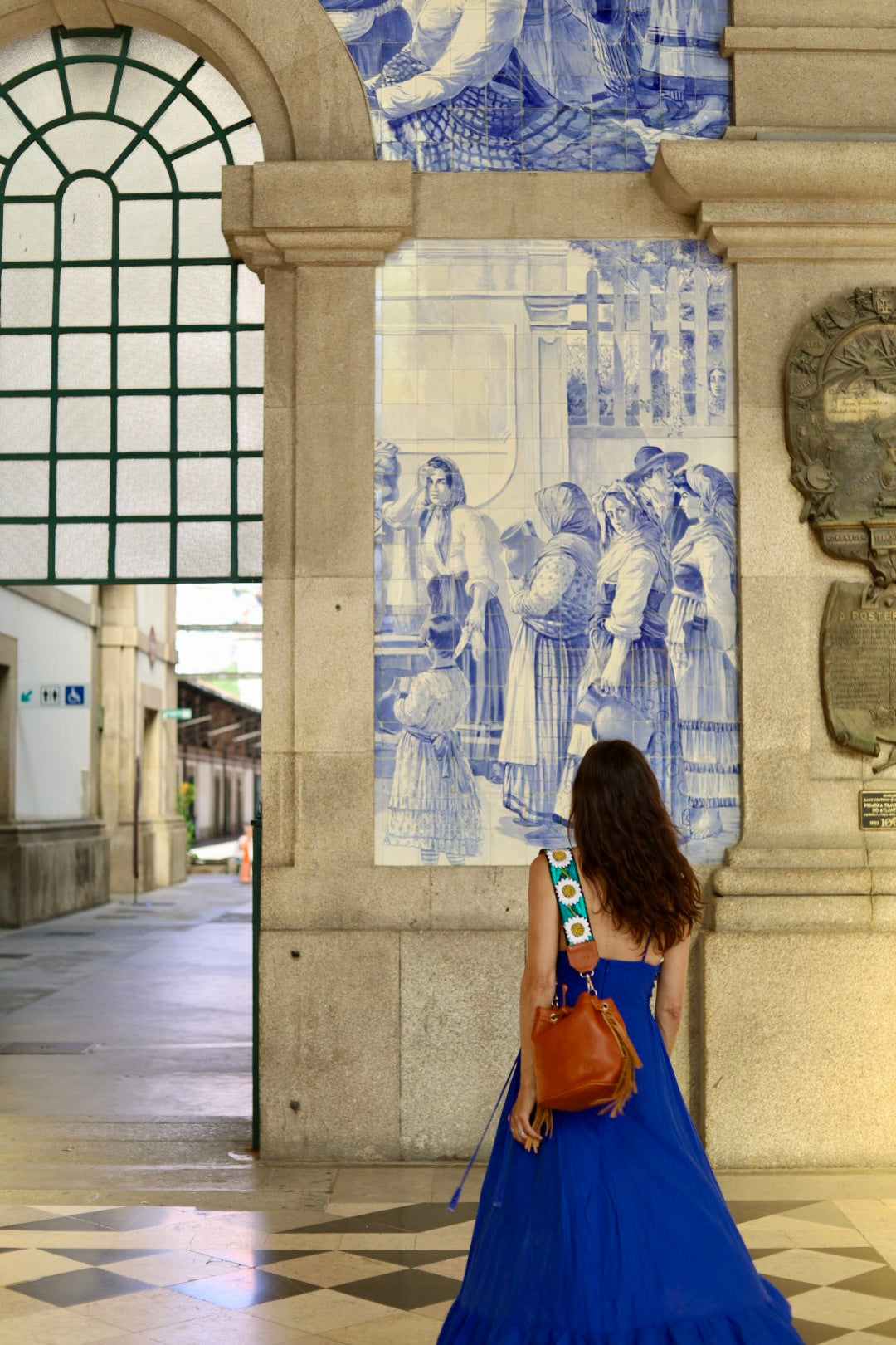A woman in a blue dress stands in front of a large blue and white ceramic tile mural inside a stone building, carrying a brown shoulder bag.