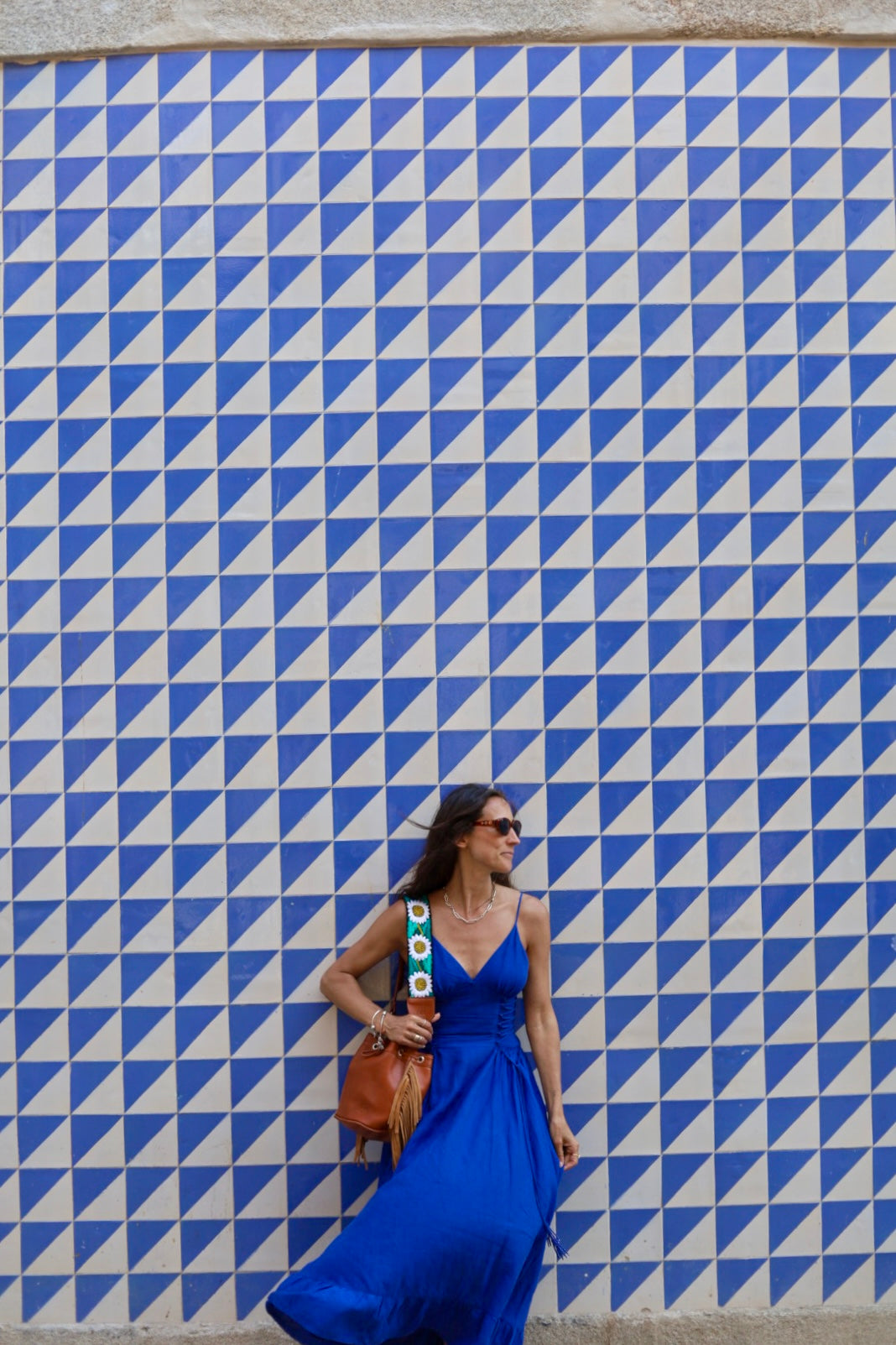 A woman in a vibrant blue sleeveless maxi dress walking against a blue and white geometric tiled wall, carrying a brown leather shoulder bag.