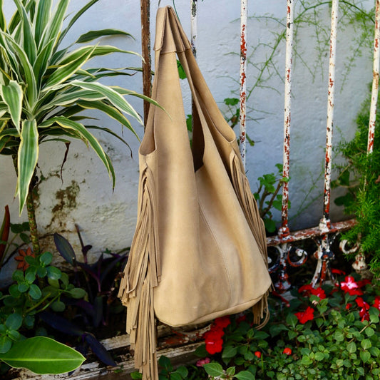 Beige leather bag with fringe hanging on a metal gate with plants and flowers in the background