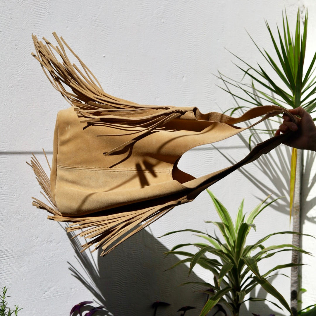 Beige handbag with fringes held against a white wall with plants in the foreground
