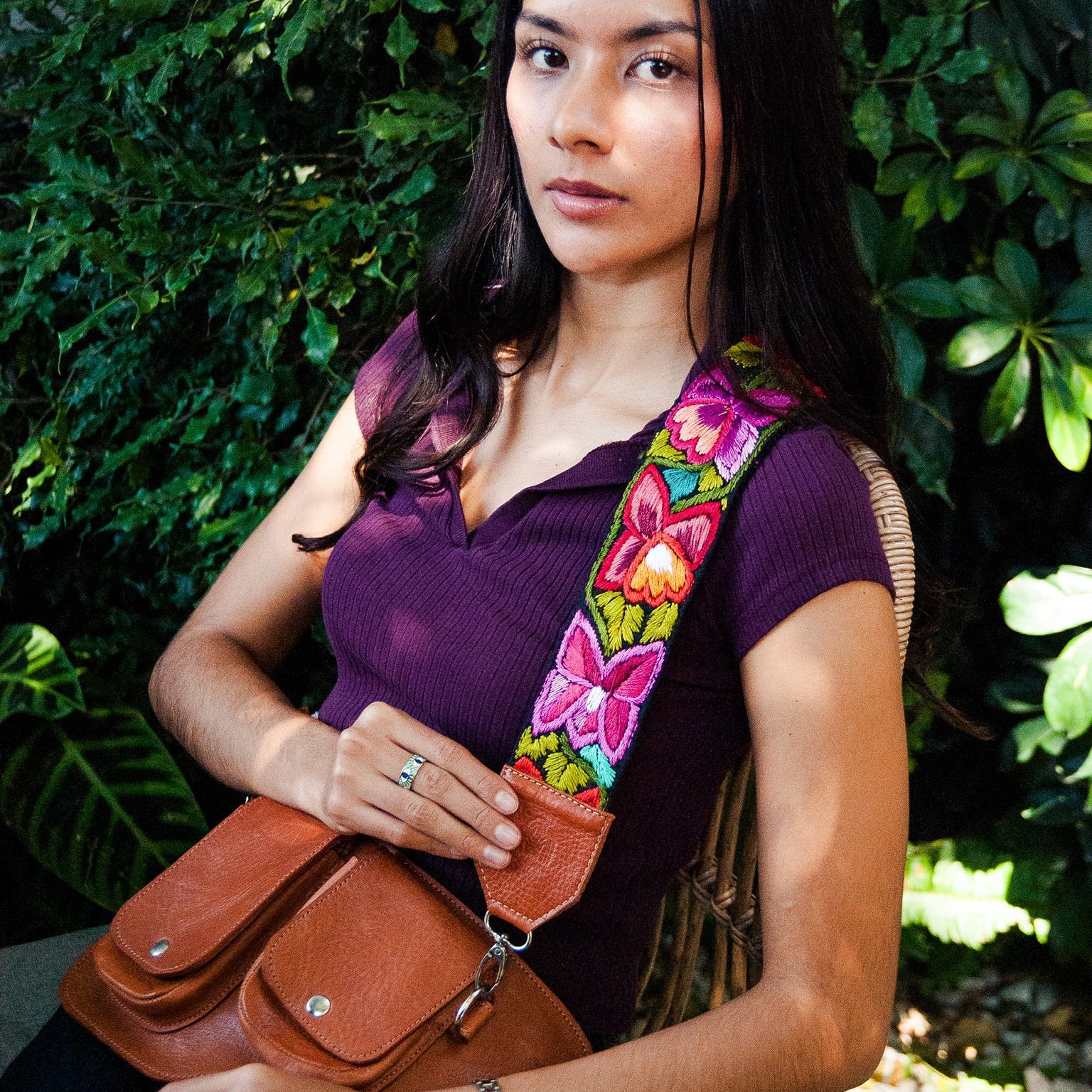Woman holding a brown leather bag with a colorful butterfly strap against a green leafy background