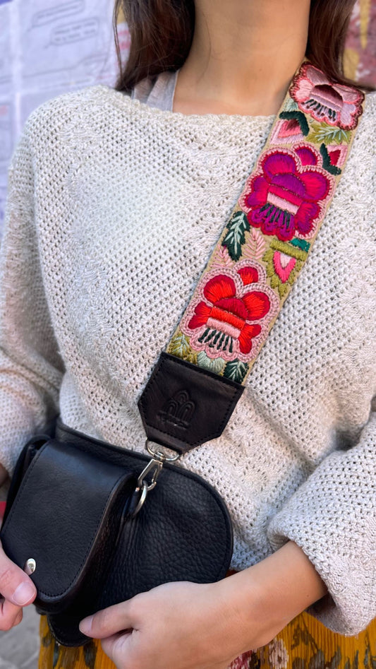 A close-up of a person wearing a cream-colored knit sweater and carrying a black leather crossbody bag with a wide, vibrant floral embroidered strap.
