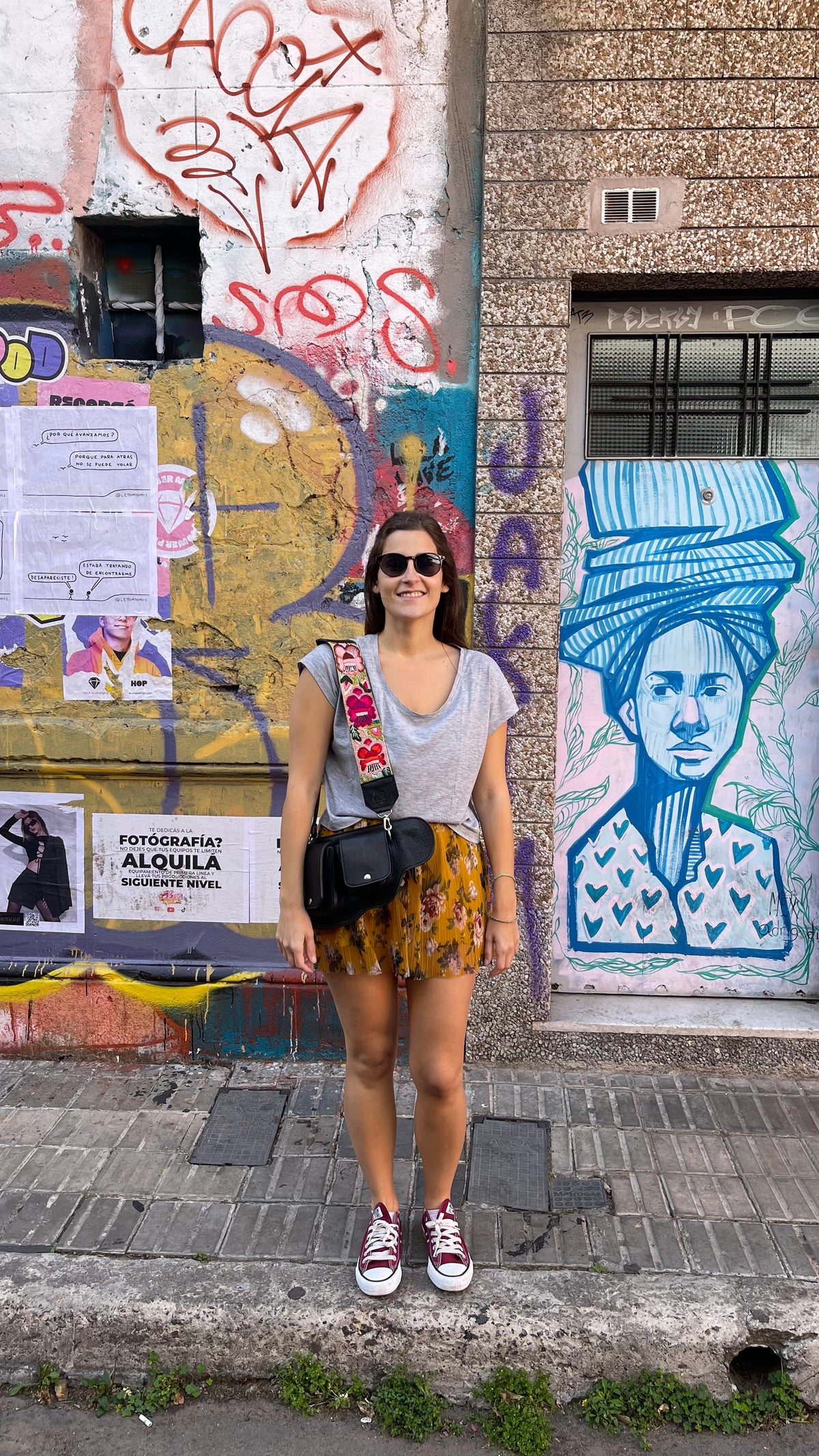 A woman wearing a gray sleeveless top, a yellow and orange patterned mini skirt, sunglasses, and red sneakers stands on a city sidewalk in front of a graffiti-covered wall.
