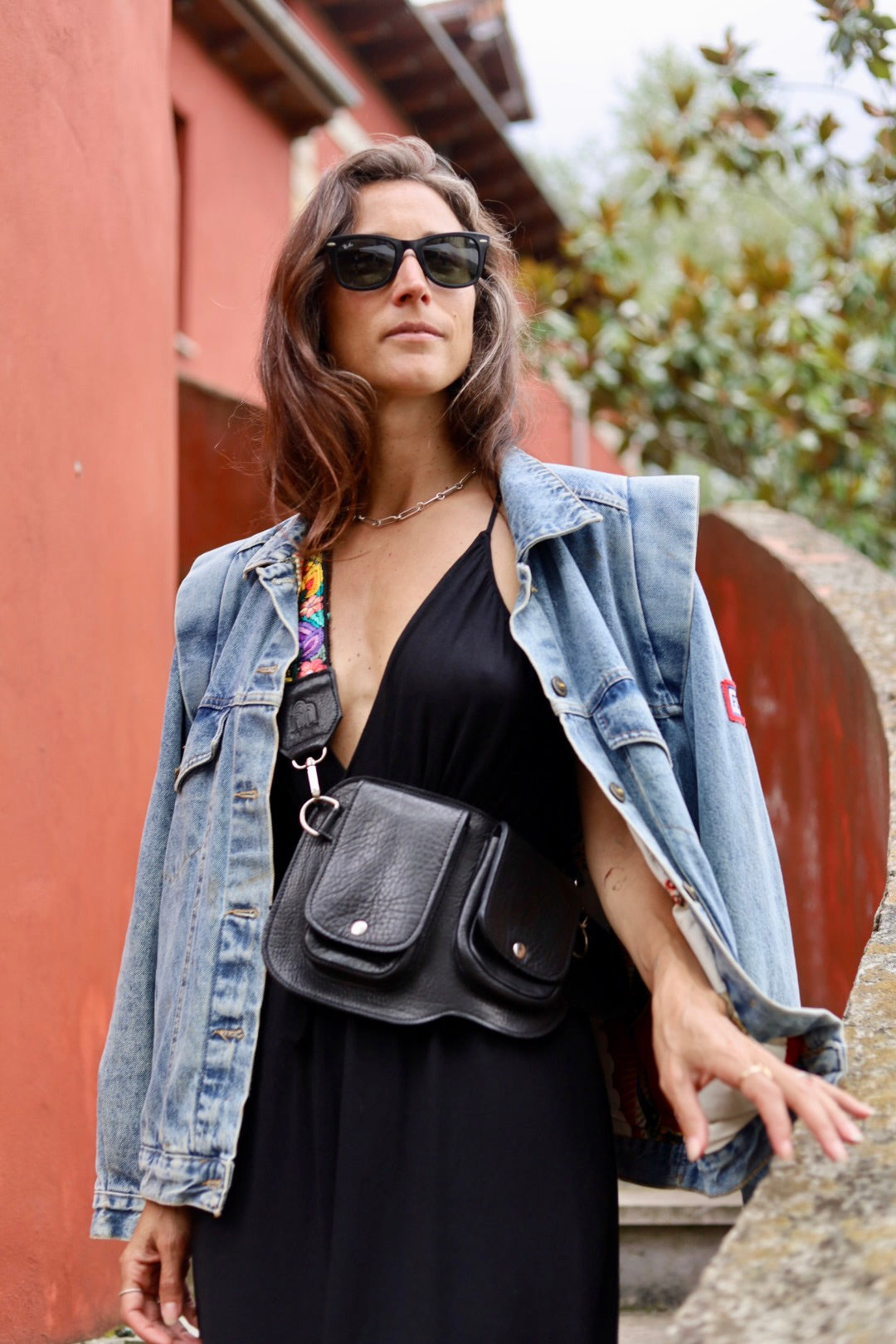 A woman wearing a black dress, a light-wash denim jacket, and black sunglasses, carrying a black leather crossbody bag with a colorful patterned strap, set against a rustic red wall and stone staircase.