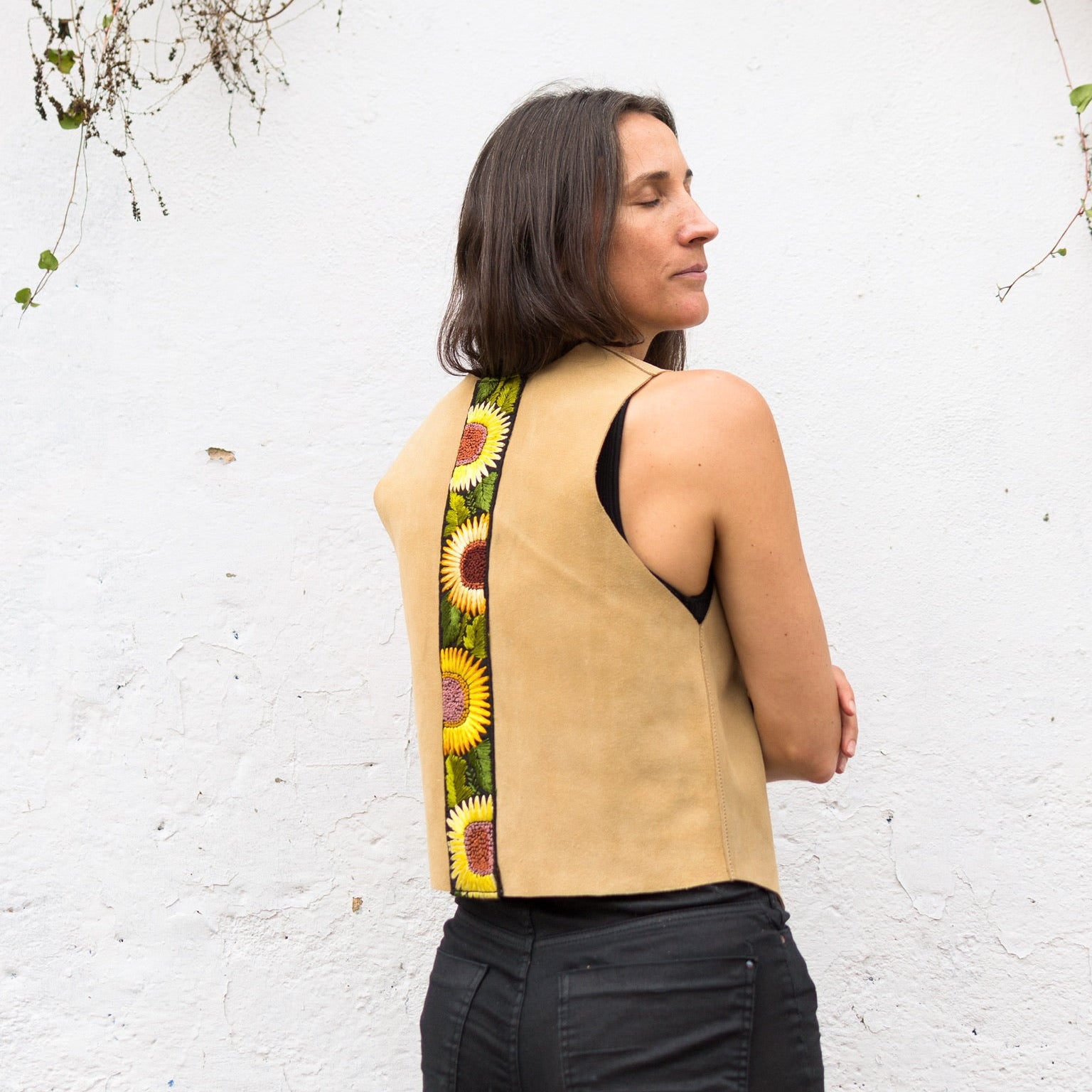 Woman wearing a beige sleeveless top with a sunflower pattern on the back, standing against a white wall with greenery.