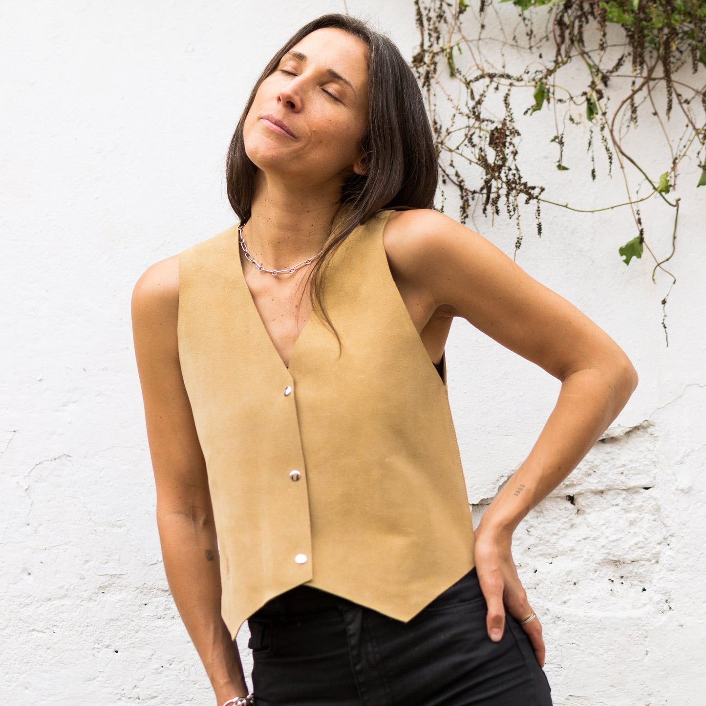 Woman wearing a beige sleeveless top and black pants against a white wall with greenery.