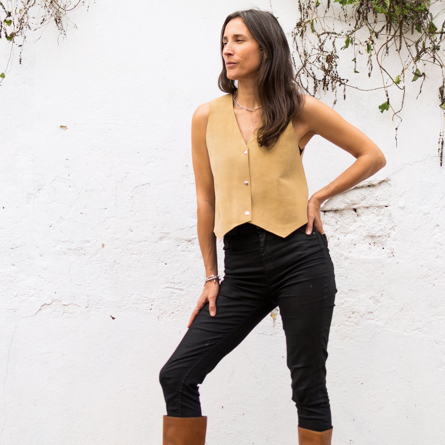 Woman wearing a beige sleeveless top, black pants, and brown boots against a white wall with greenery.
