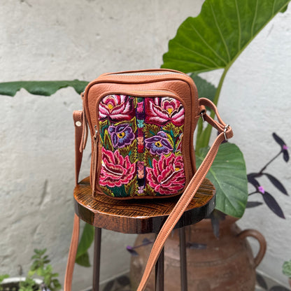 Colorful embroidered bag with a brown leather strap on a small wooden stool against a textured wall.