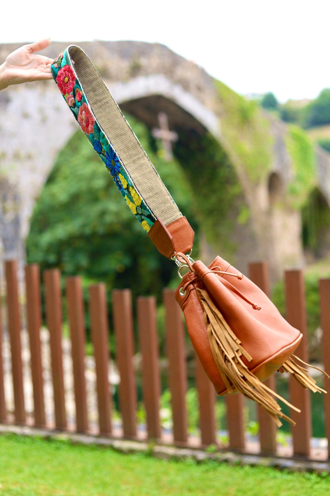 A tan leather bucket bag with a wide, colorful floral embroidered shoulder strap, held up by a person's hand against a blurred outdoor background of a stone archway and wooden fence.