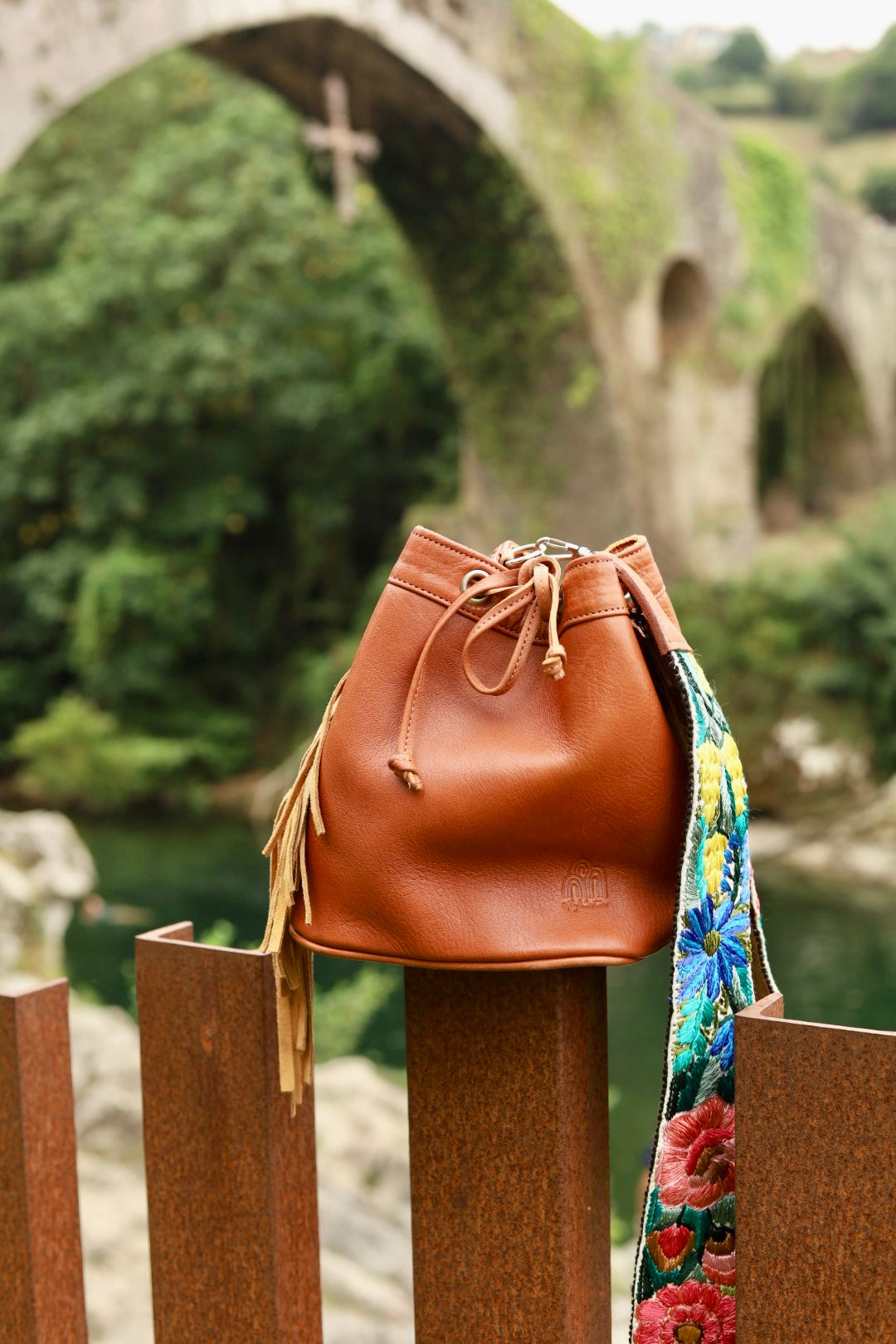 A brown leather bucket bag with a colorful floral printed strap, resting on a rustic wooden fence in front of a blurred stone bridge and green landscape.