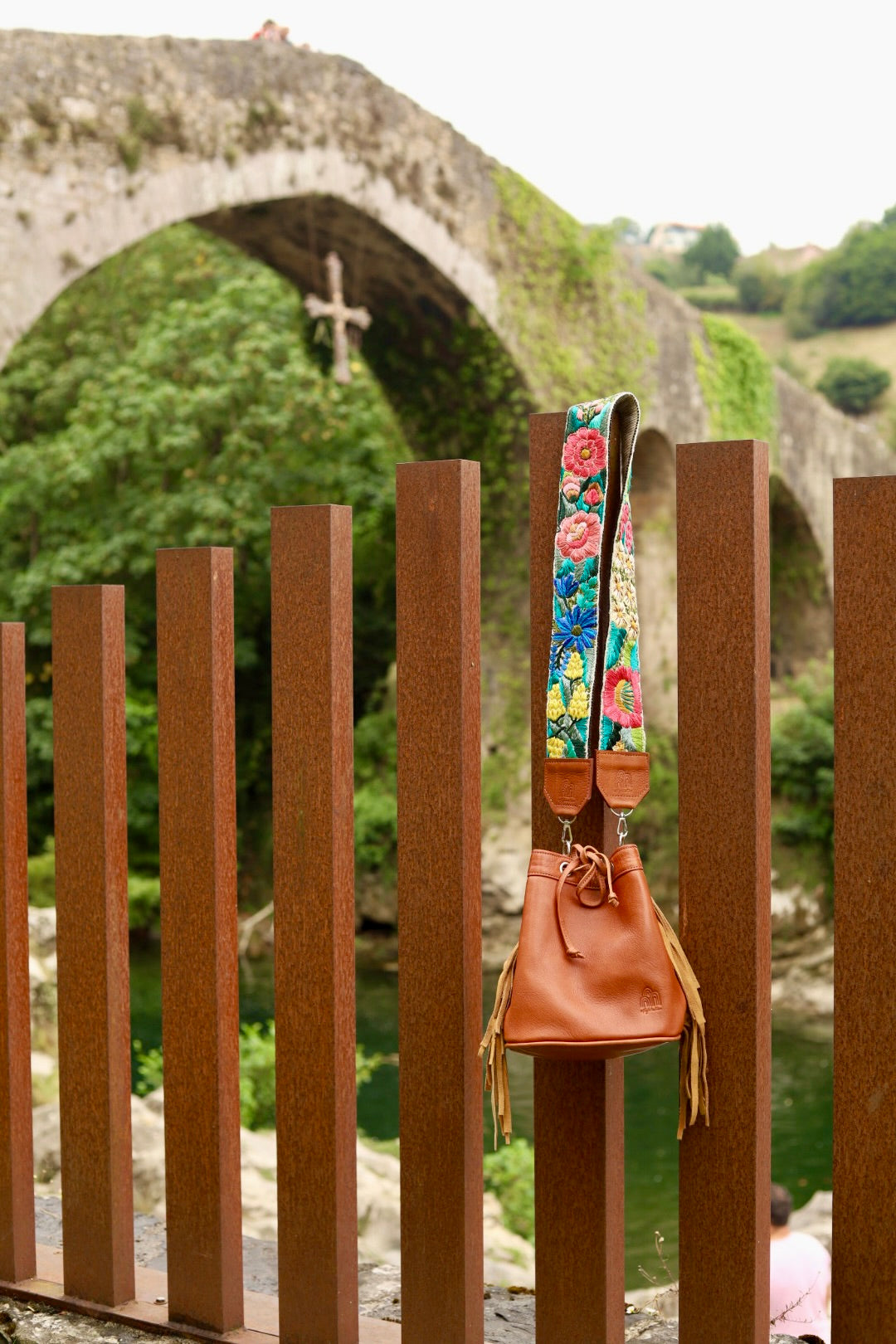 A brown leather bucket bag with a colorful floral patterned shoulder strap hangs from a rusty brown metal fence in front of a stone archway over green water.
