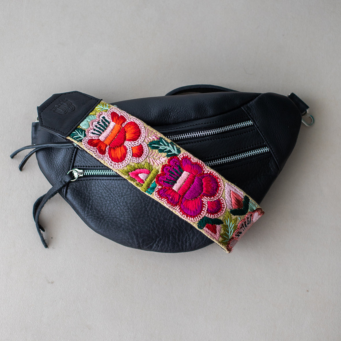 Black sling bag with a floral patterned strap, laid flat on a grey surface.