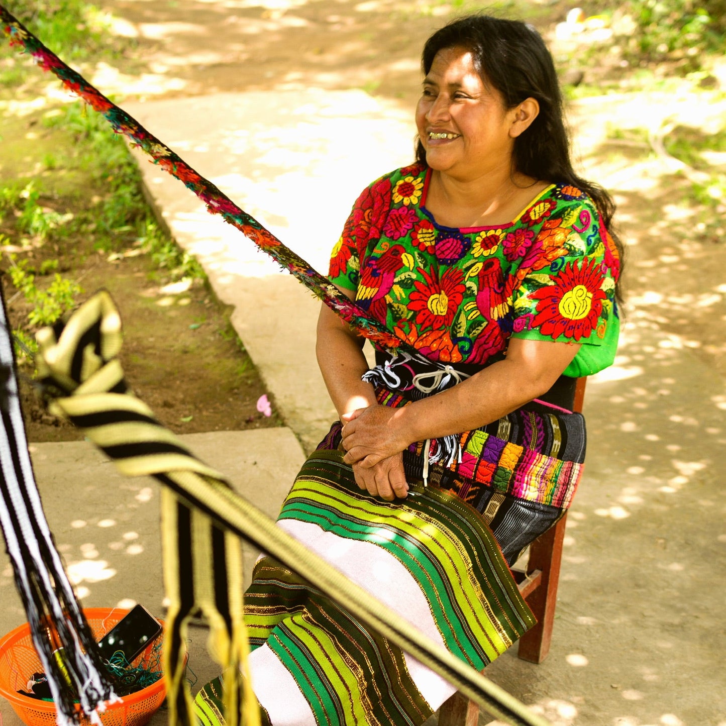 Woman in colorful traditional clothing sitting on a wooden chair with a loom in front of her.