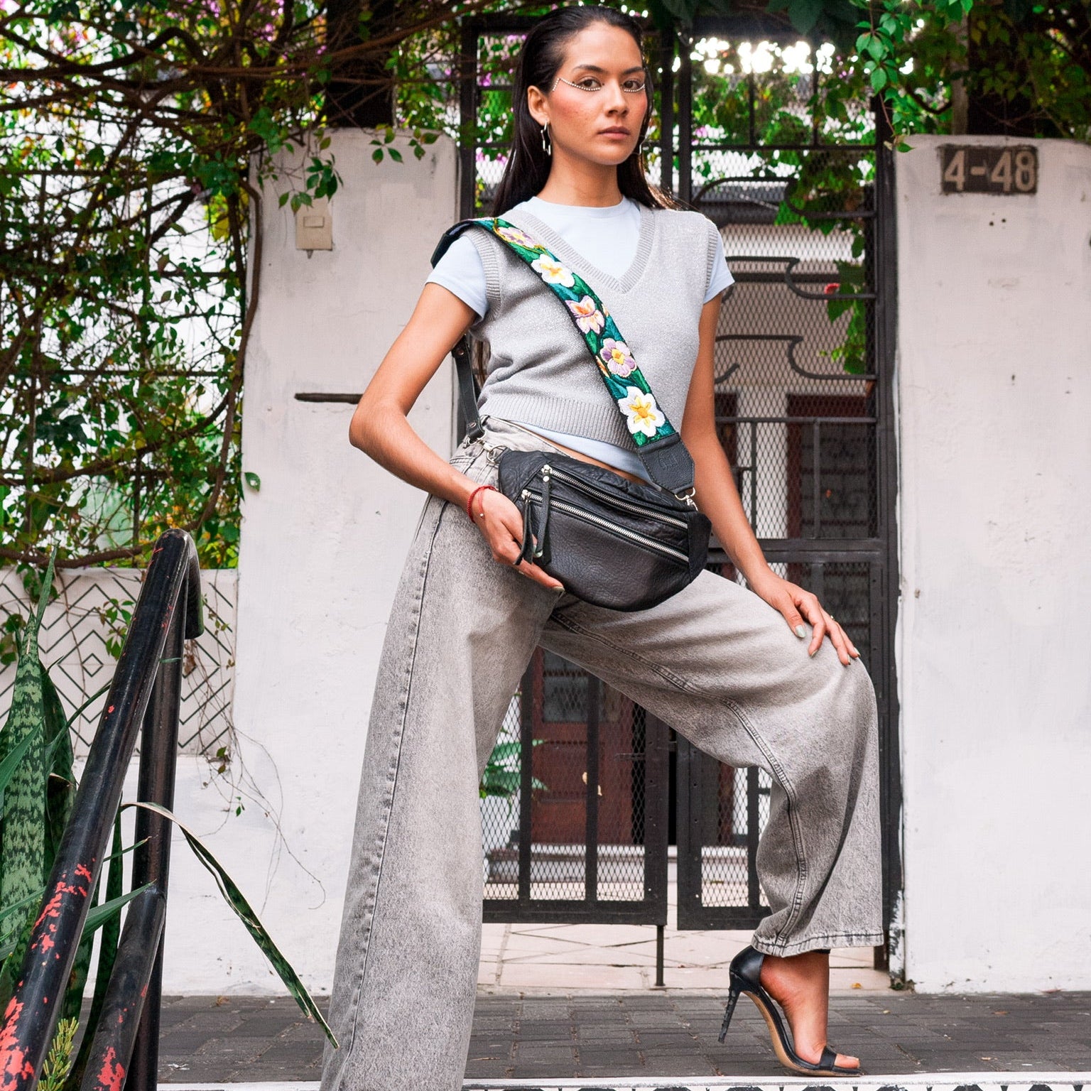 Woman holding a black handbag in an outdoor setting with greenery and a white wall.