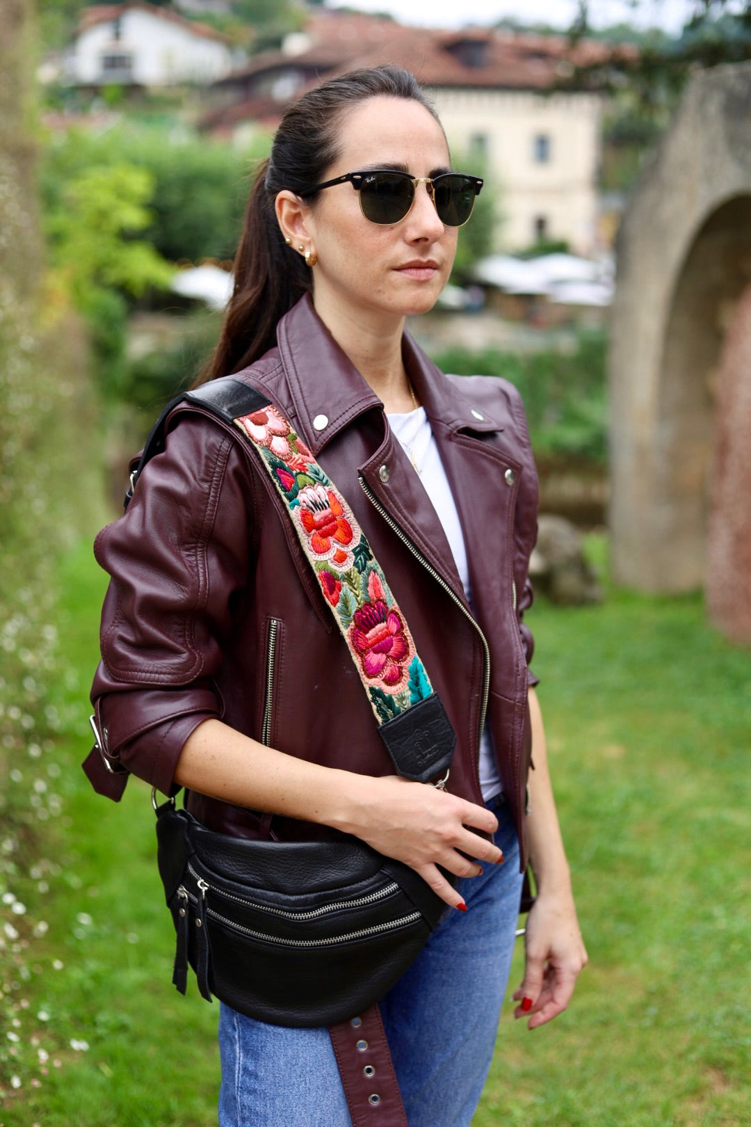 A woman wearing a dark burgundy leather moto jacket with a colorful floral embroidered shoulder strap, blue jeans, and black sunglasses, carrying a black crossbody fanny pack outdoors.