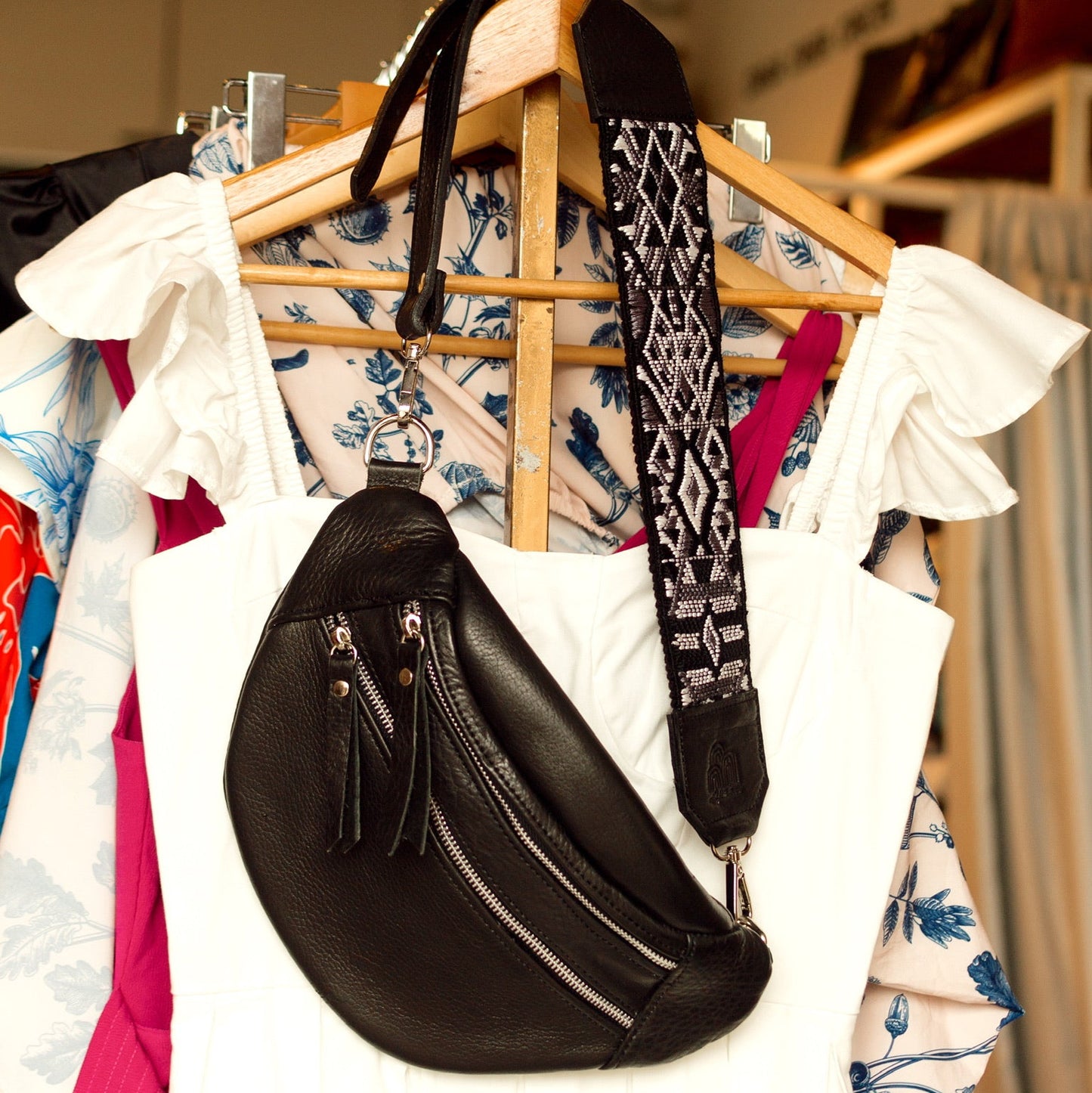 Black leather bag with patterned strap on a clothing rack with colorful garments.