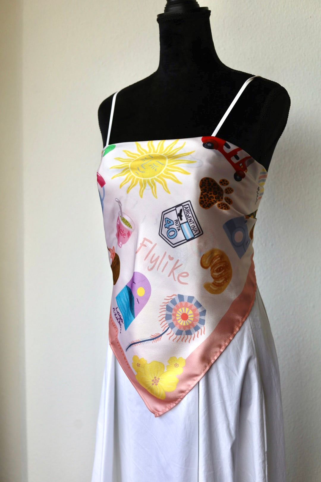 A white satin camisole top with a colorful graphic print featuring a sun, balloons, a paw print, and the word 'Flylike', displayed on a black dress form mannequin.