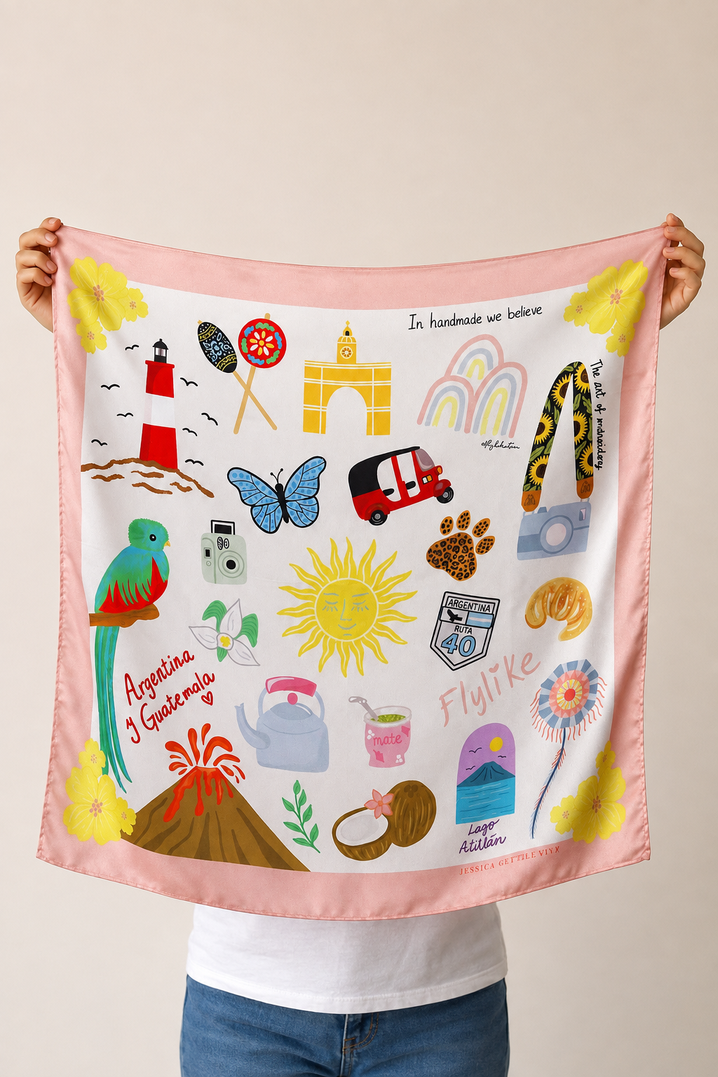 A person holds up a square pink scarf featuring colorful travel-themed illustrations, including a lighthouse, parrot, sun, rainbow, and small icons, against a plain beige background.