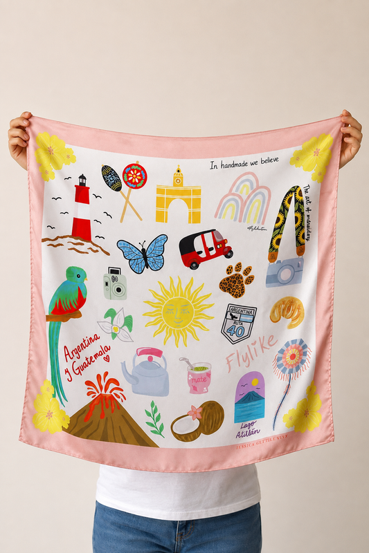 A person holds up a square pink scarf featuring colorful travel-themed illustrations, including a lighthouse, parrot, sun, rainbow, and small icons, against a plain beige background.