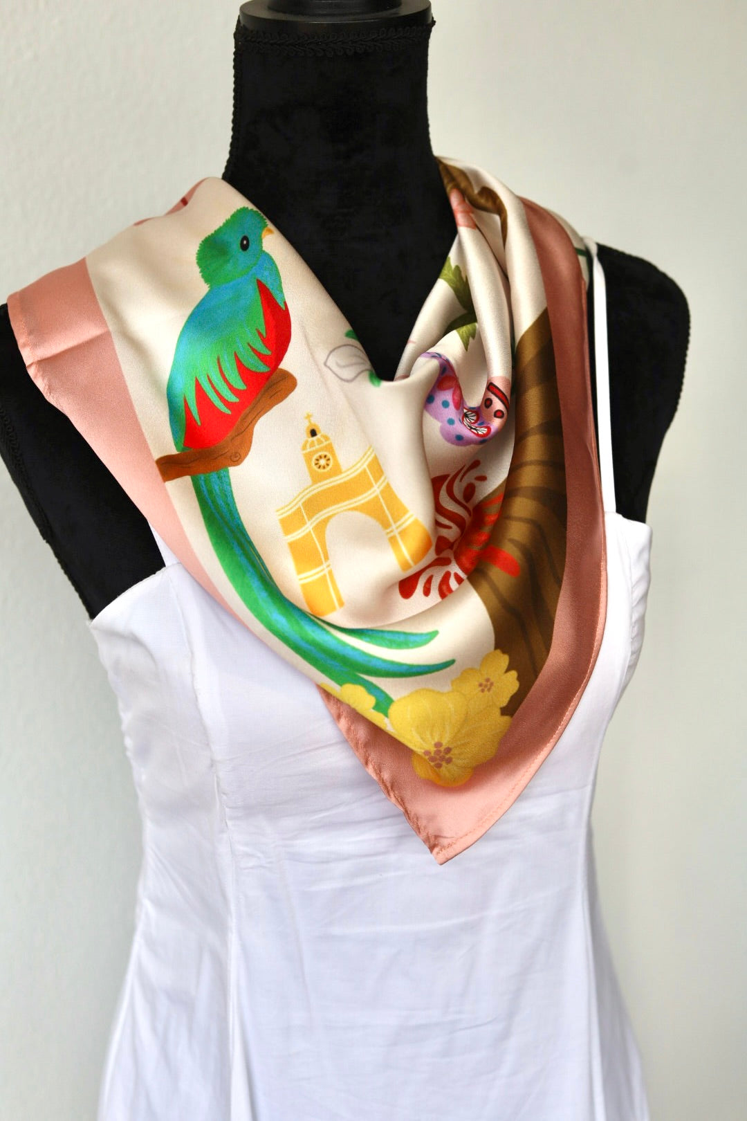 A square silk scarf with a colorful print of a green and red quetzal bird, a golden arch, and a pink border, draped on a black mannequin torso wearing a white top.